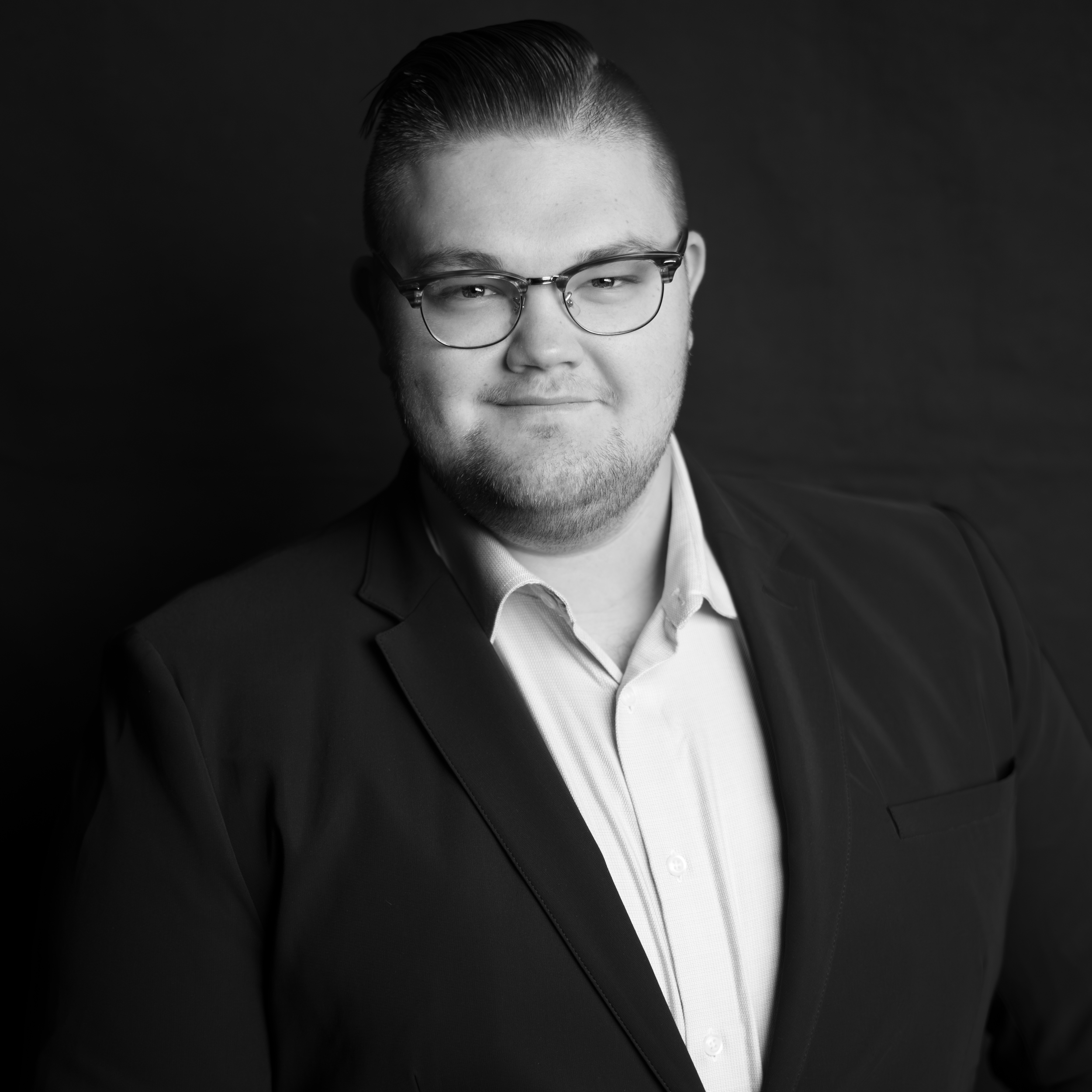 Portrait of a young man with glasses, short hair, light beard, wearing a suit jacket and collared shirt, smiling subtly, against a dark background.