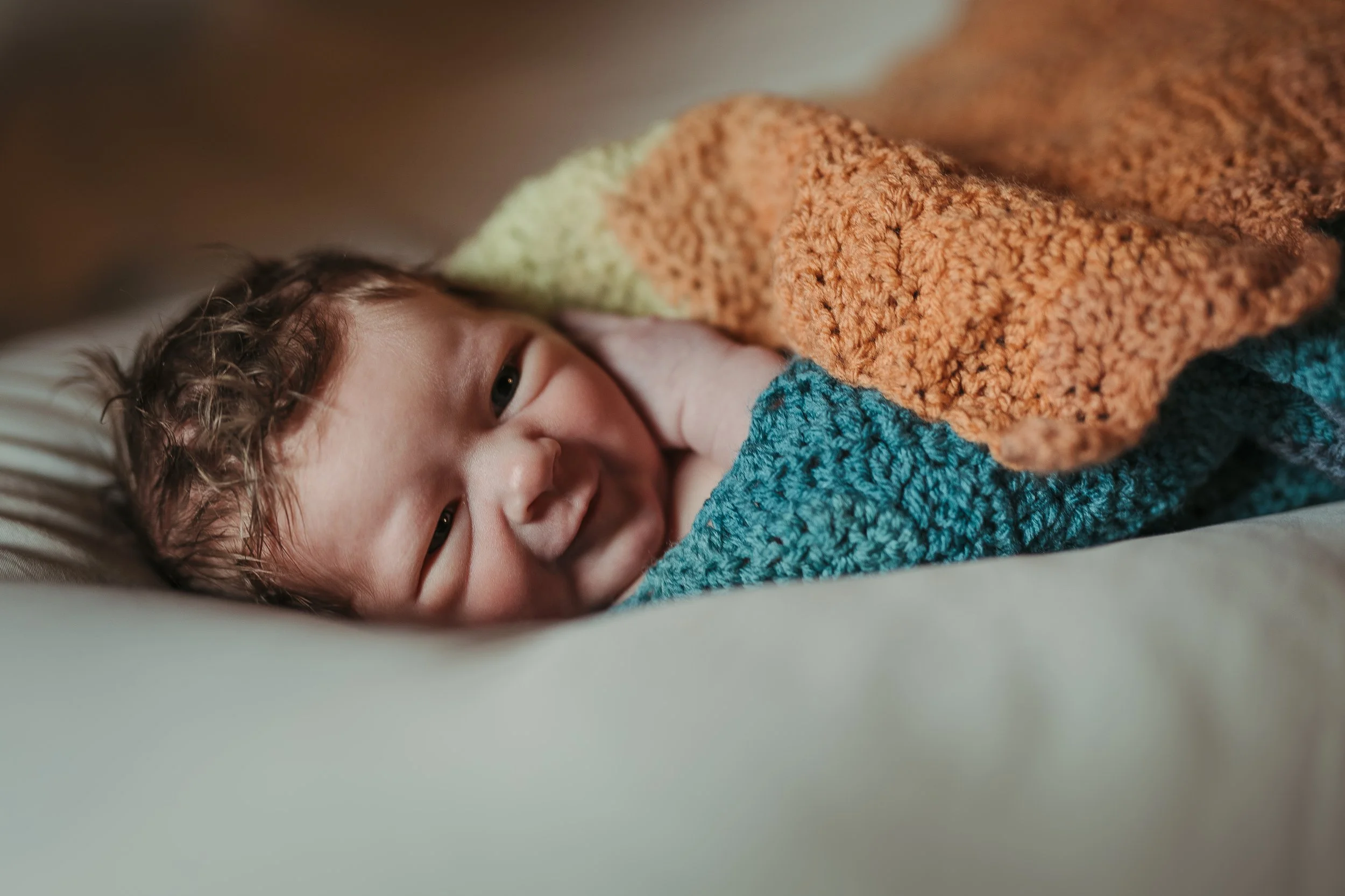 A smiling baby with curly hair lying on a bed, partially covered with a multicolored crocheted blanket.