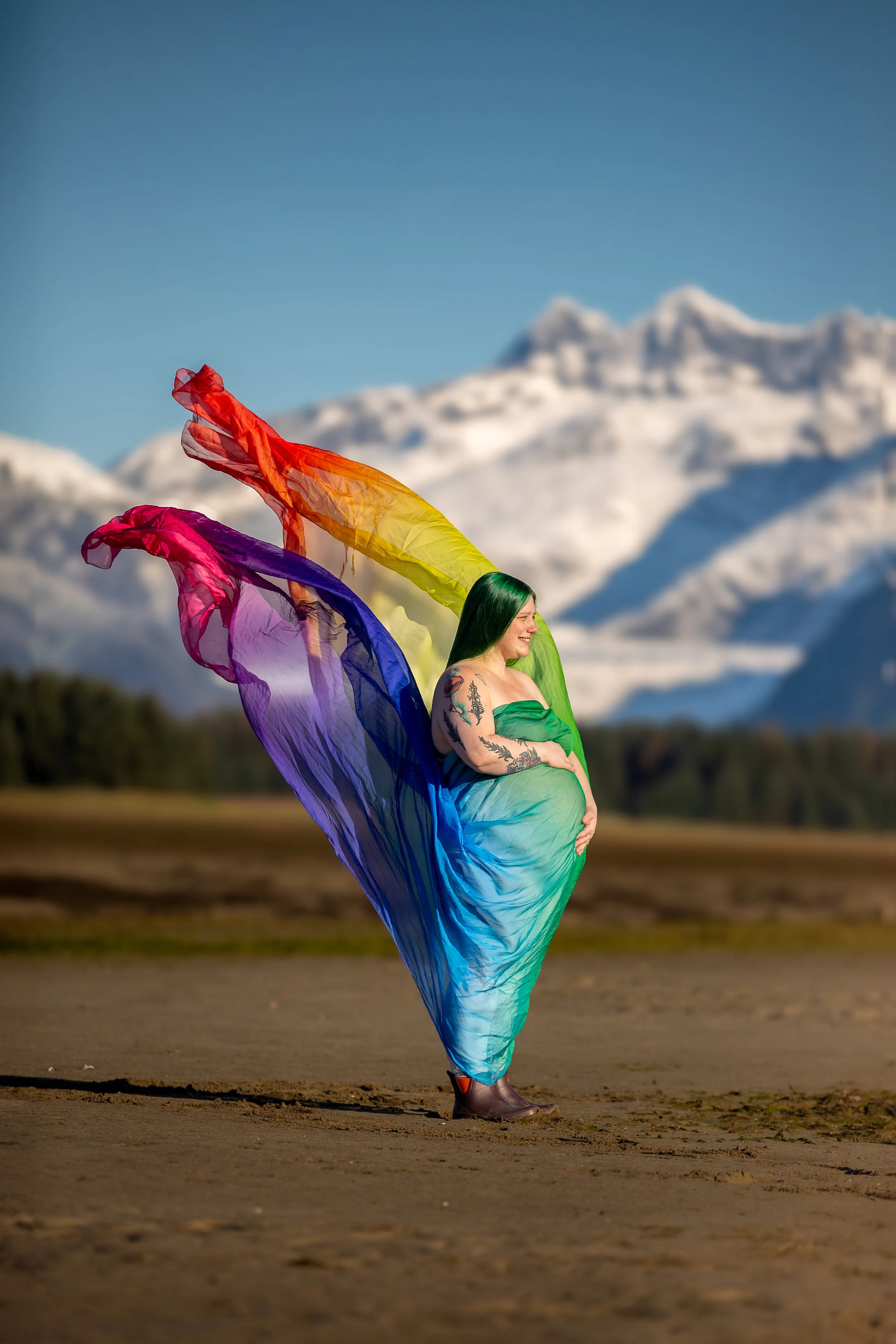 Andria (white woman with green hair) pregnant wrapped in a rainbow silk and standing in front of a glacier