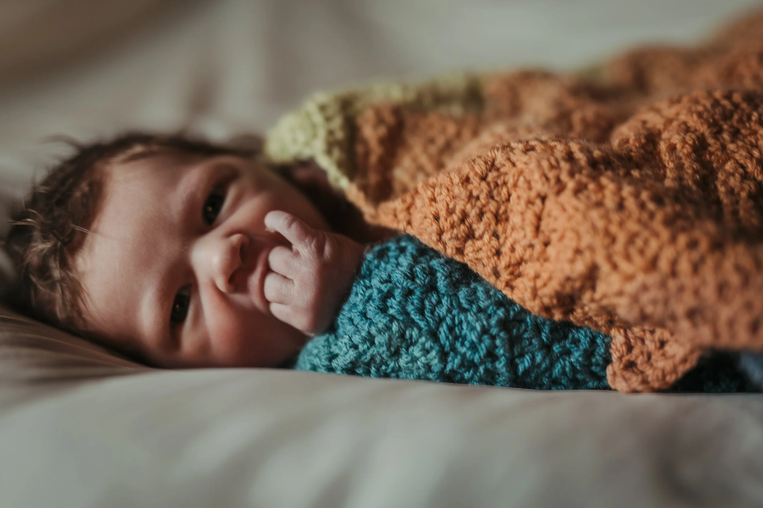 Close-up of a baby lying on a bed, with one hand near their mouth, wrapped in colorful crocheted blankets.