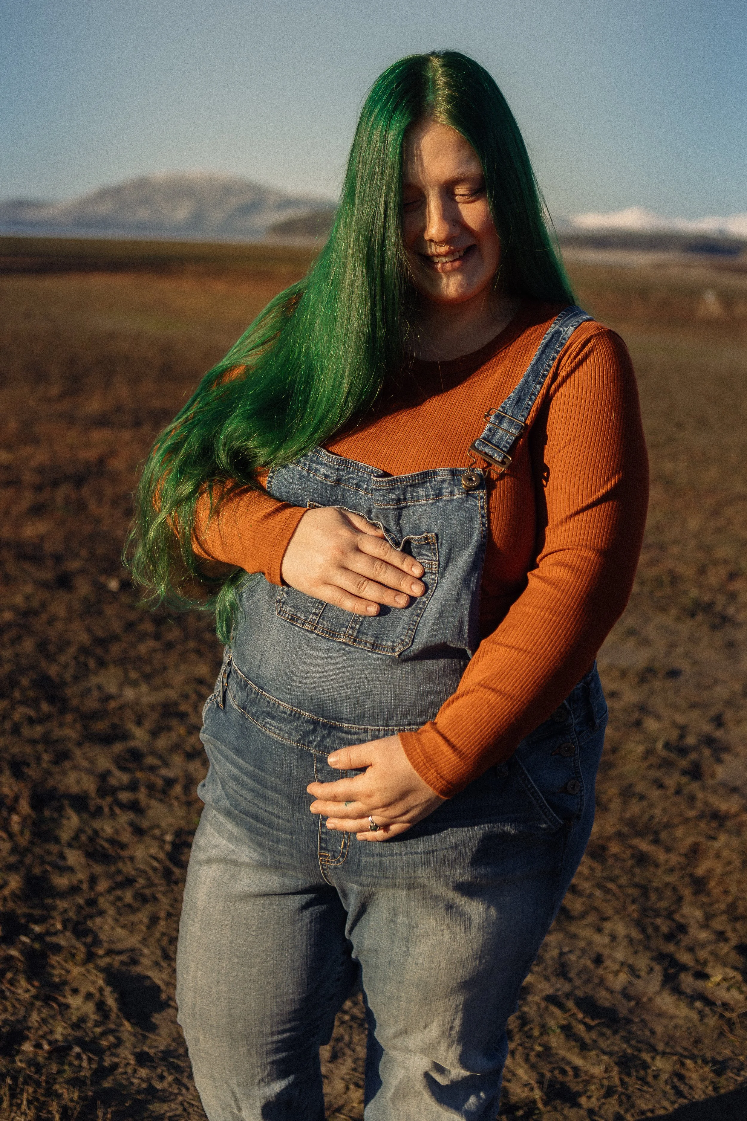 Andria (white woman with green hair) holding her pregnant belly during an outdoor maternity photoshoot