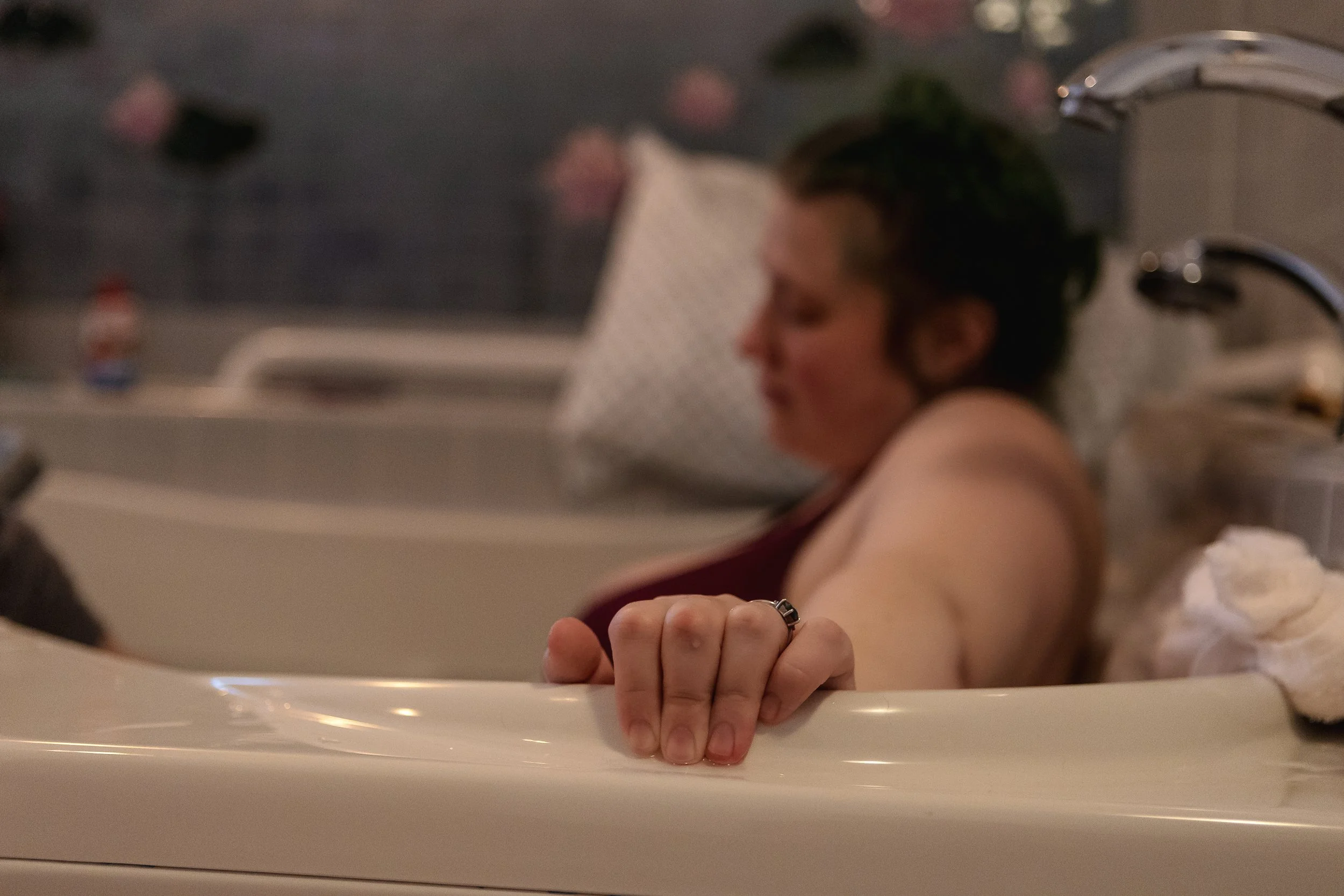 A woman with dark hair in a birthing tub, leaning back with her arm resting on the edge, wearing a dark top, with a blurred background.