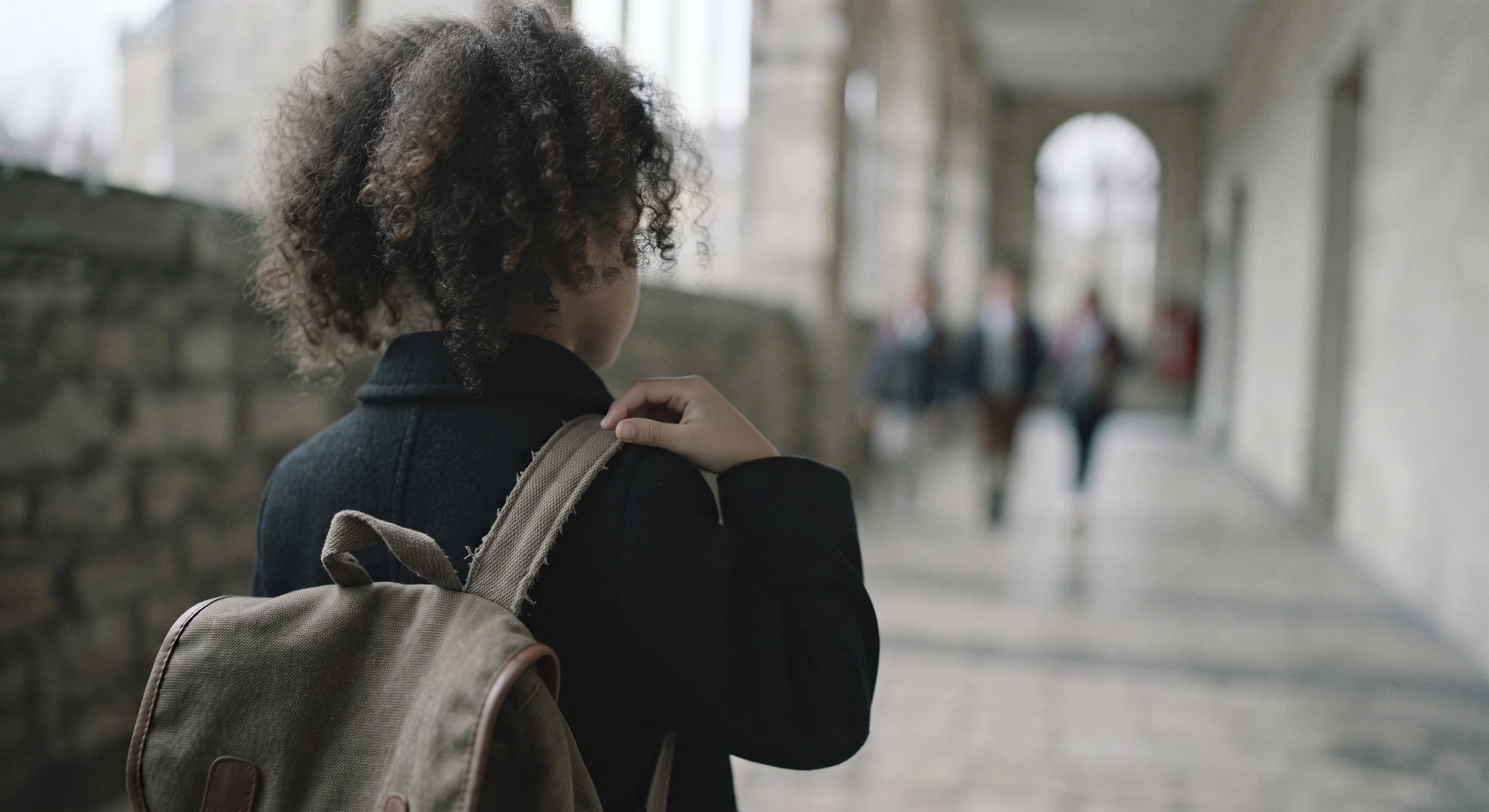 Une jeune fille avec des cheveux bouclés portant un sac à dos beige dans un couloir avec des arches en pierre et des personnes en arrière-plan.