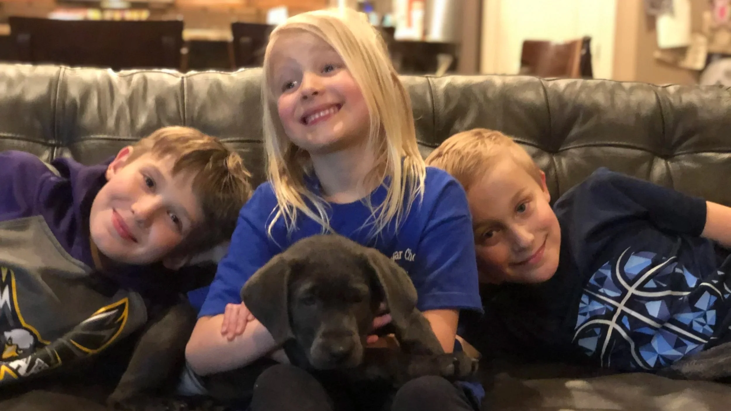 Three kids and a puppy lying on a leather couch smiling at the camera in a cozy indoor setting.