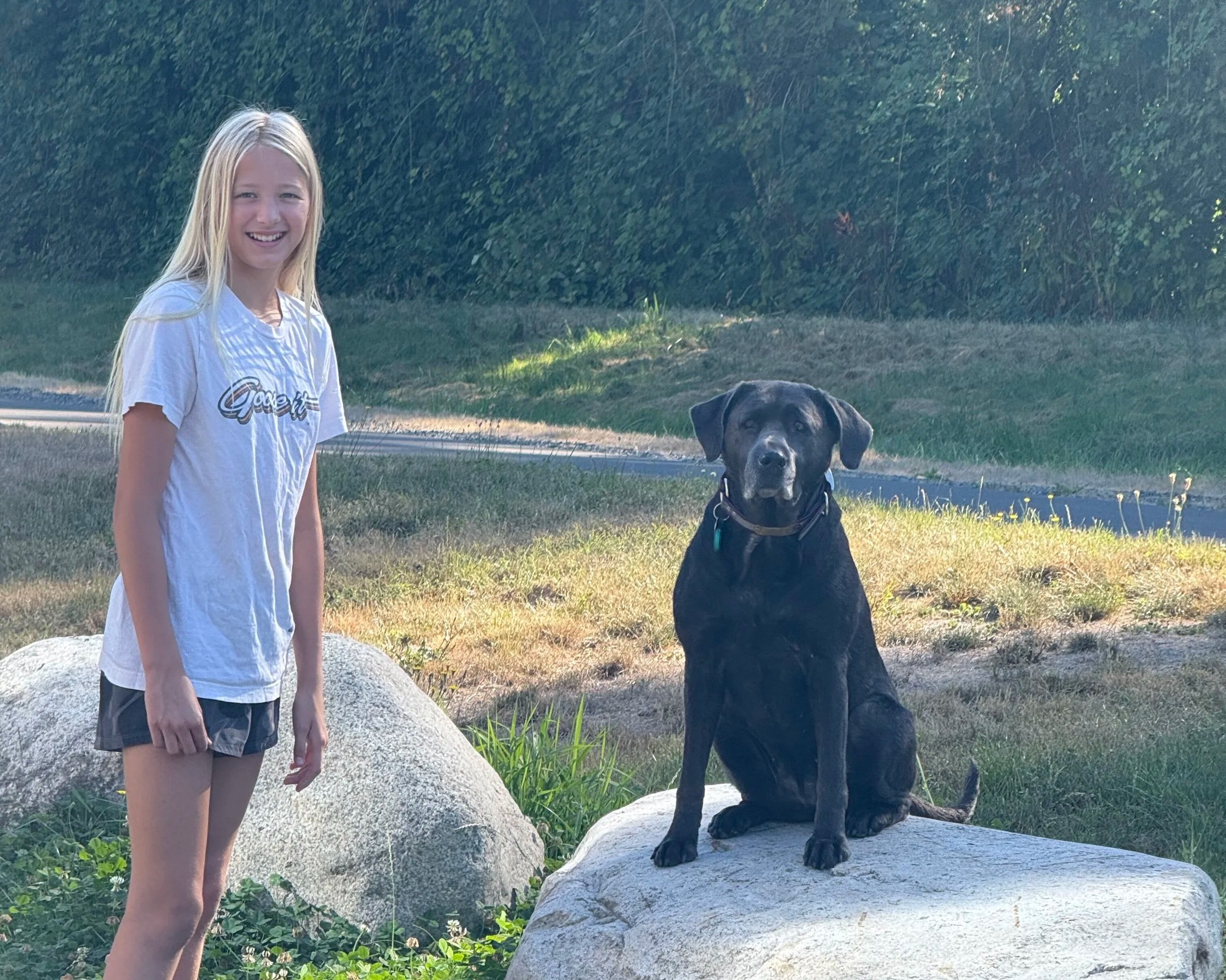 A girl with long blonde hair smiling and standing next to a black dog sitting on a large rock in a grassy area with trees in the background, during daytime.
