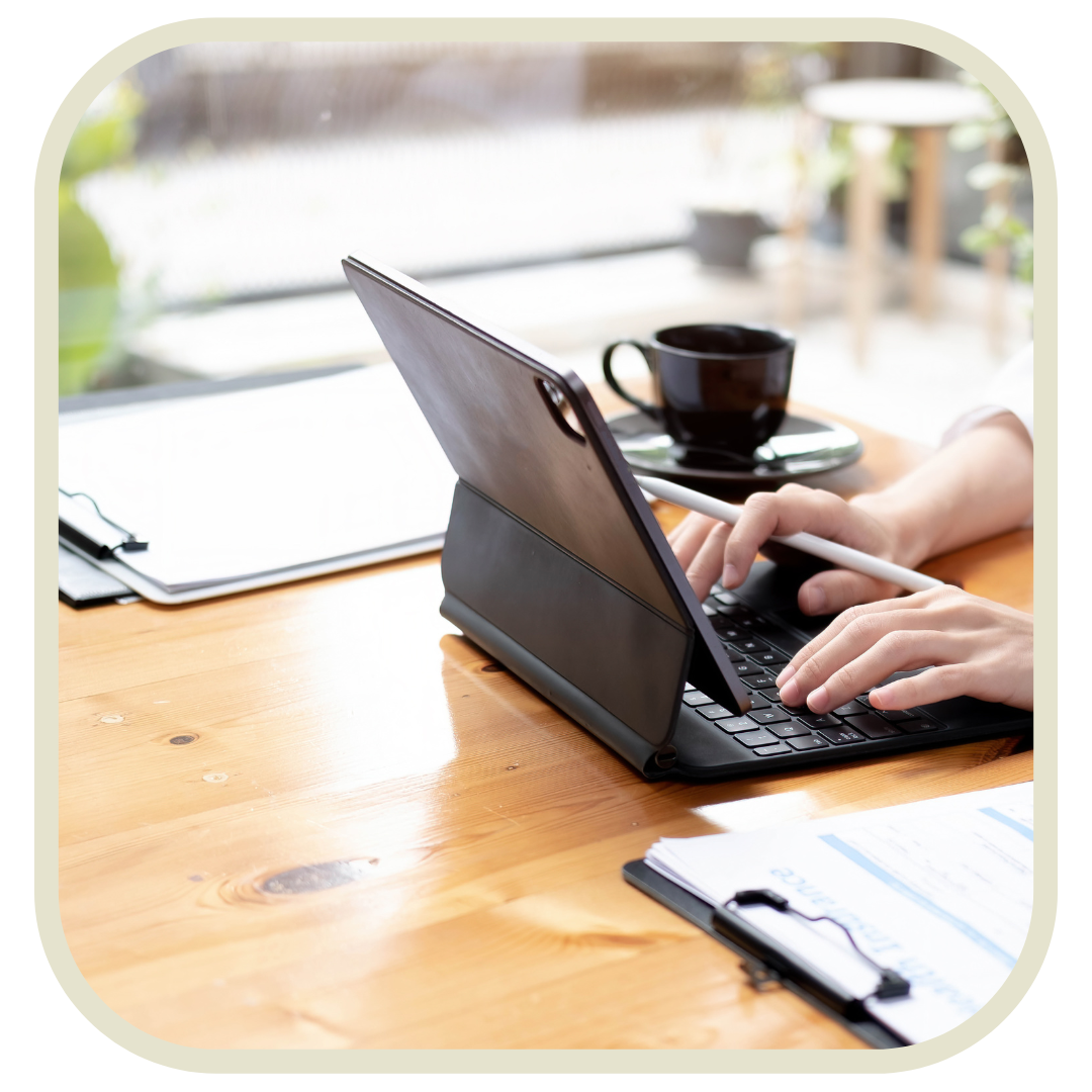 Person working on a tablet with a keyboard and stylus at a wooden desk, with a black coffee cup and saucer, some papers, and a clipboard nearby in a bright indoor setting.