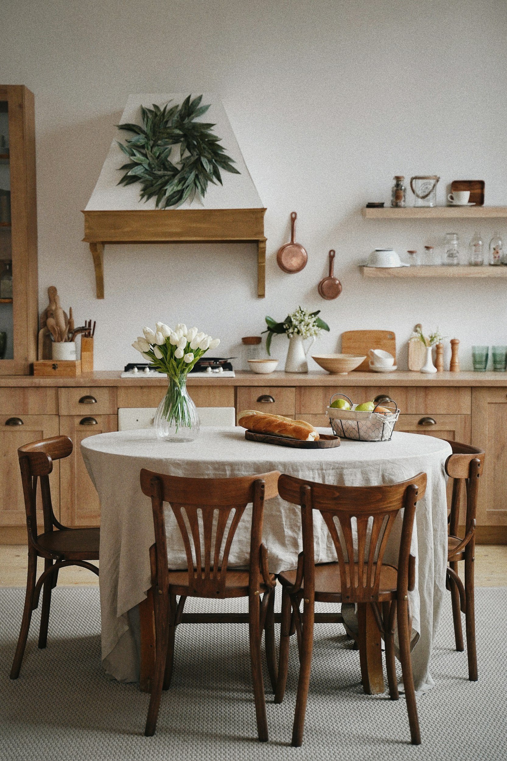 A cozy kitchen with a round dining table covered with a beige tablecloth, holding a vase of white tulips, a loaf of bread, and a basket of green apples. The kitchen features wooden cabinets, open shelves, a range hood, and copper pans hanging on the wall.