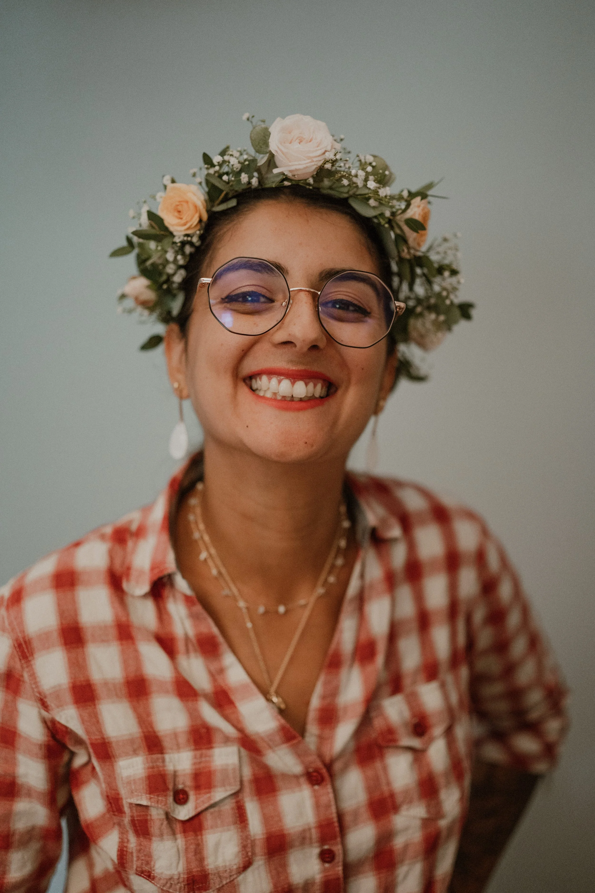 Organisatrice de mariage souriante portant une couronne de fleurs, lunettes rondes, bijoux en argent, chemise à carreaux rouge et blanc.