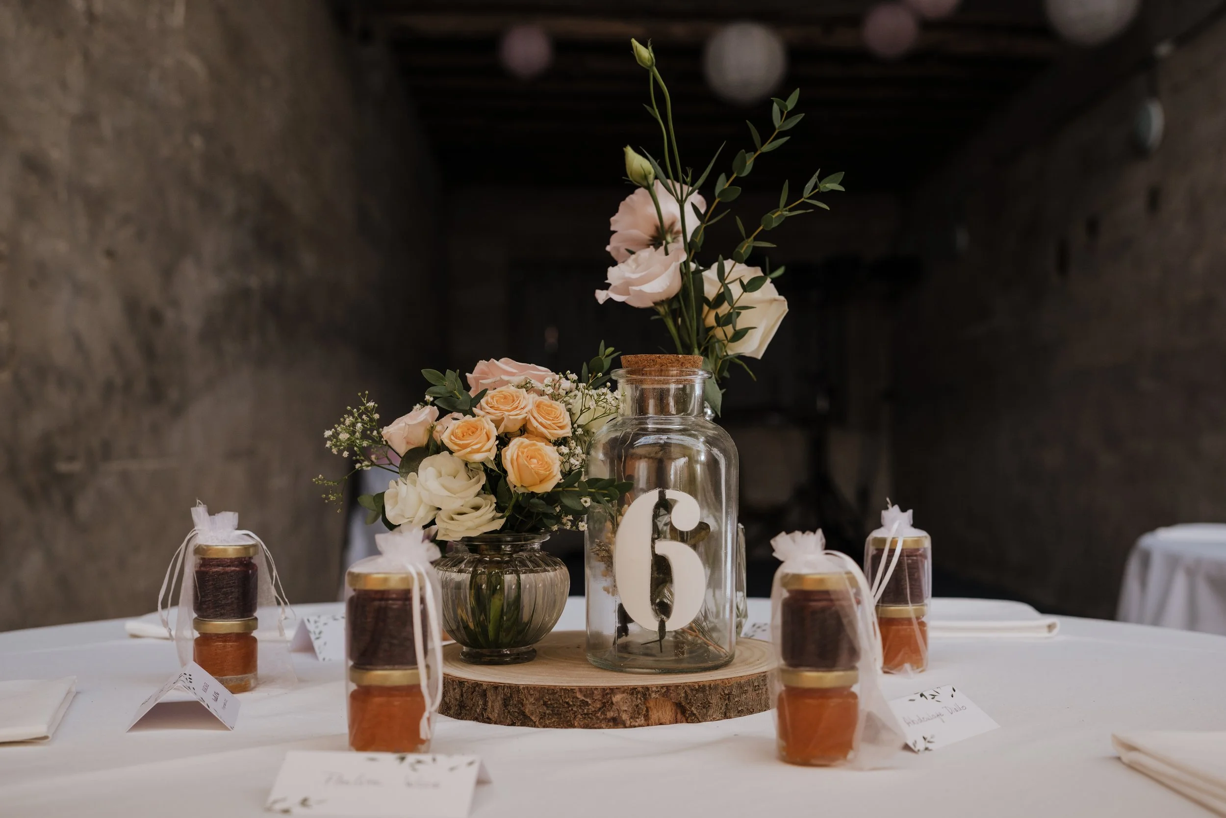 Centre de table de mariage avec fleurs roses et blanches, jars de confiture avec étiquettes, numéro 6 en blanc dessiné sur une bouteille en verre, assiettes, nappe blanche, décoration rustique dans un lieu à murs en pierre.