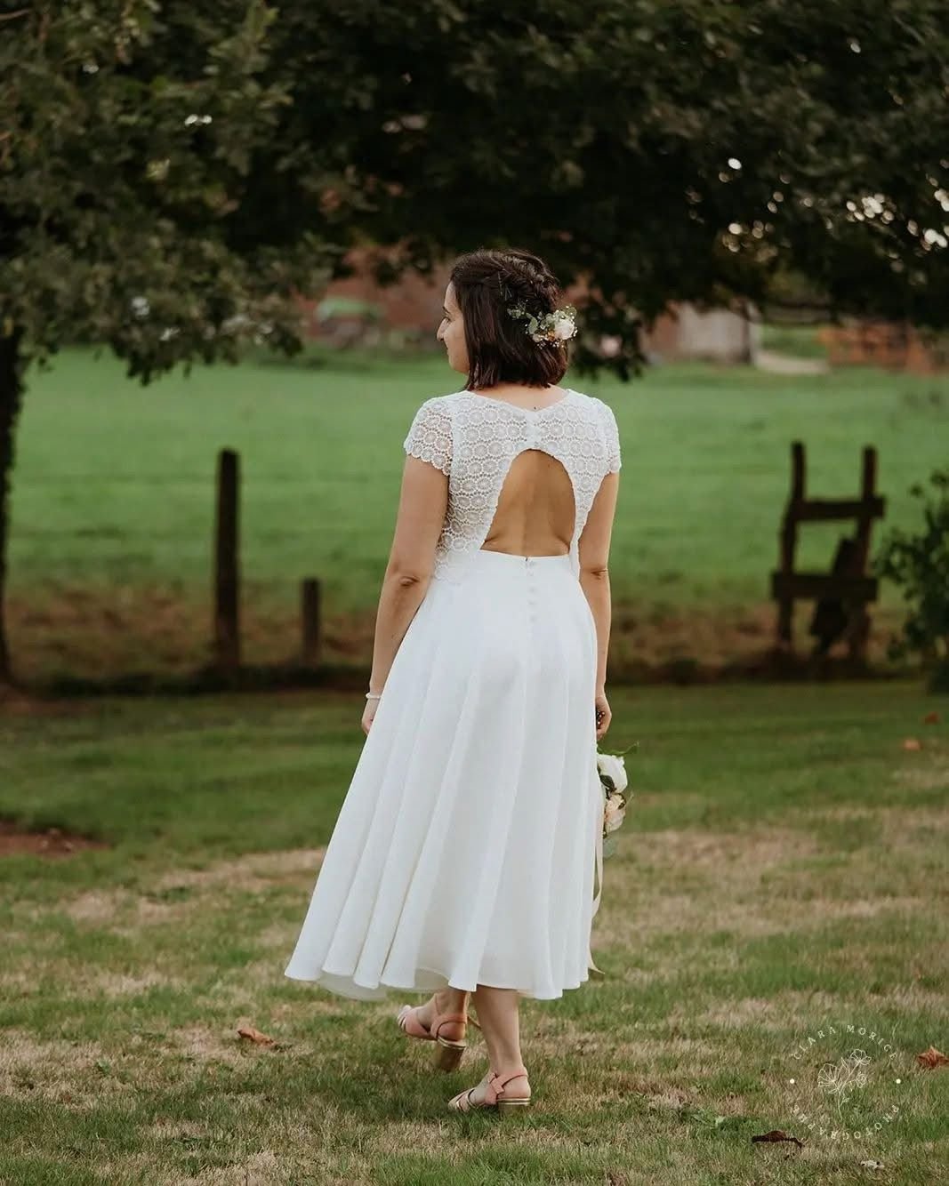 Une femme en robe blanche de mariage avec un dos nu, tenant un bouquet, dans un jardin avec des arbres et une clôture en bois.