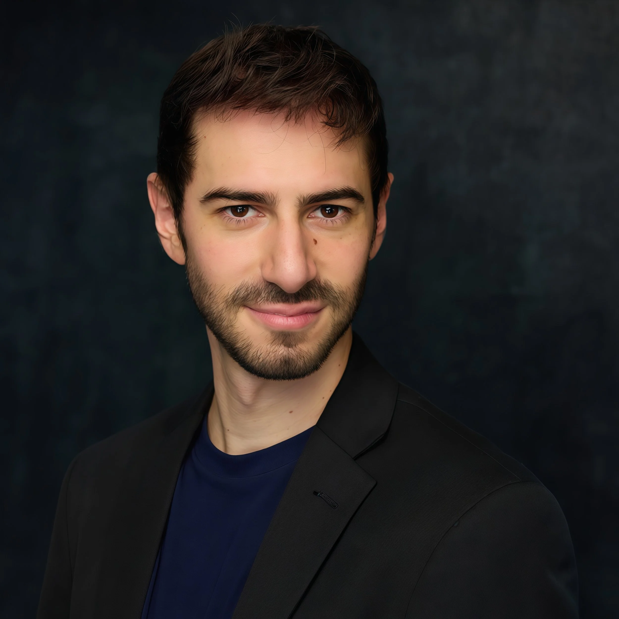A young man with dark hair and a beard, wearing a black blazer over a navy shirt, smiling slightly and looking into the camera against a dark background.
