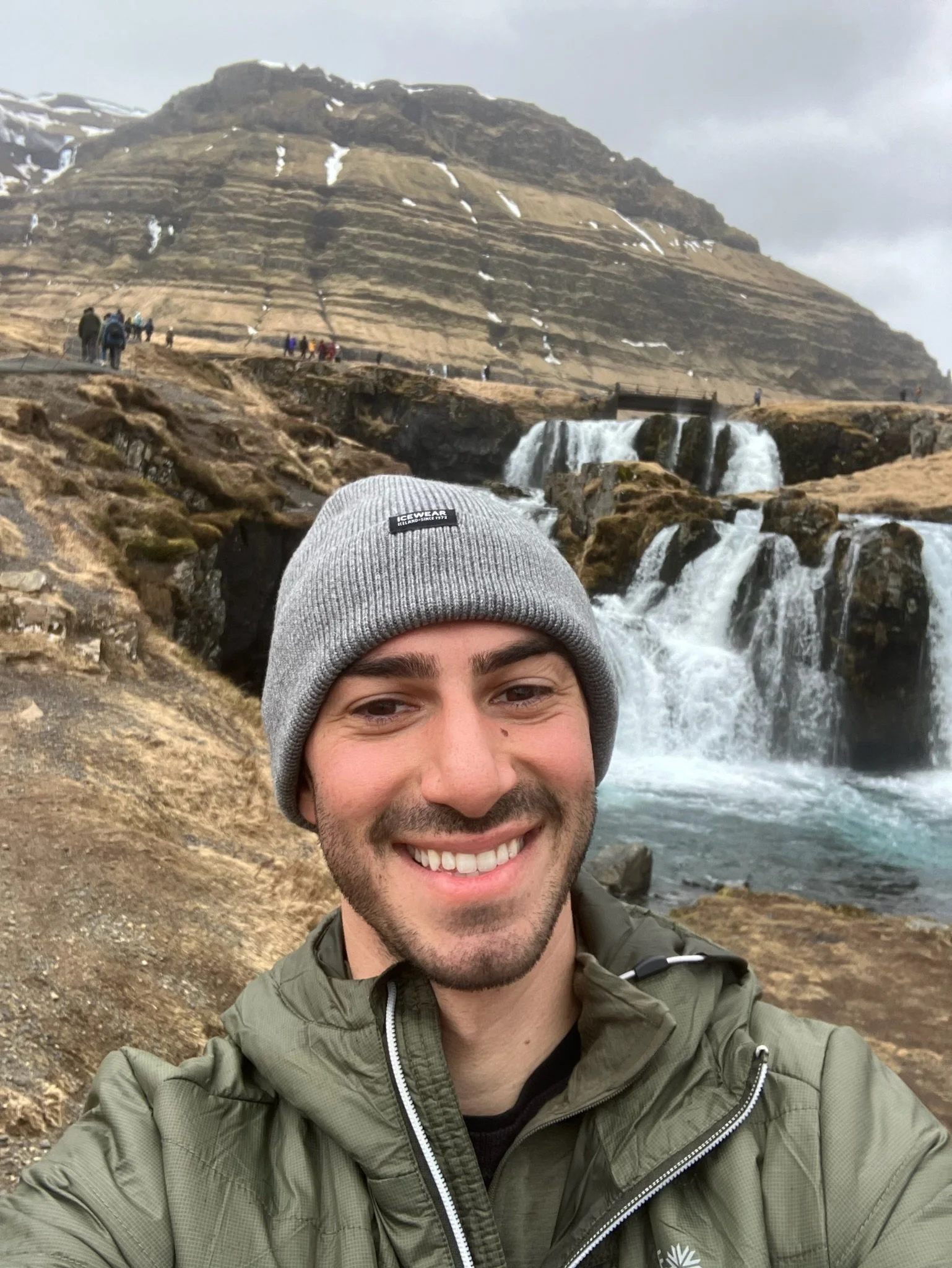 A man smiling for a selfie outdoors in front of a waterfall with multiple cascades and a rocky landscape, with a mountain in the background and a cloudy sky.