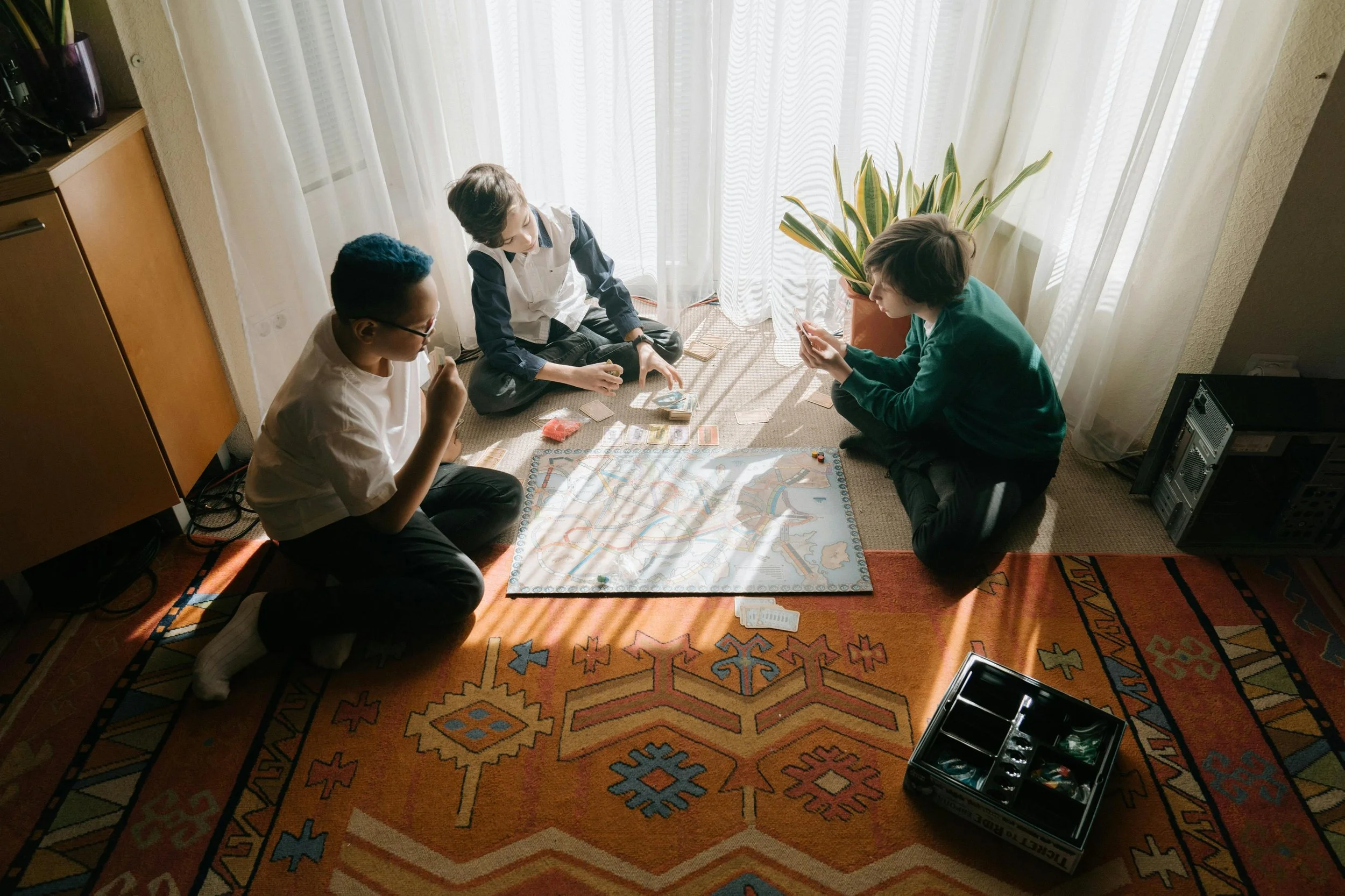 Three children playing a board game on a carpeted floor next to a window with sheer curtains, sunlight streaming in, with a potted plant nearby.