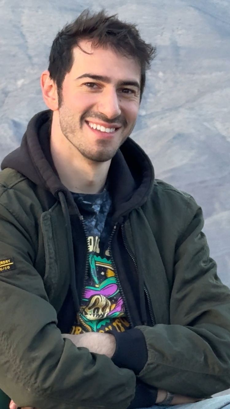 A young man with dark hair and a beard smiling at the camera, wearing a dark green jacket, a hoodie, and a colorful graphic t-shirt, with rocky terrain in the background.