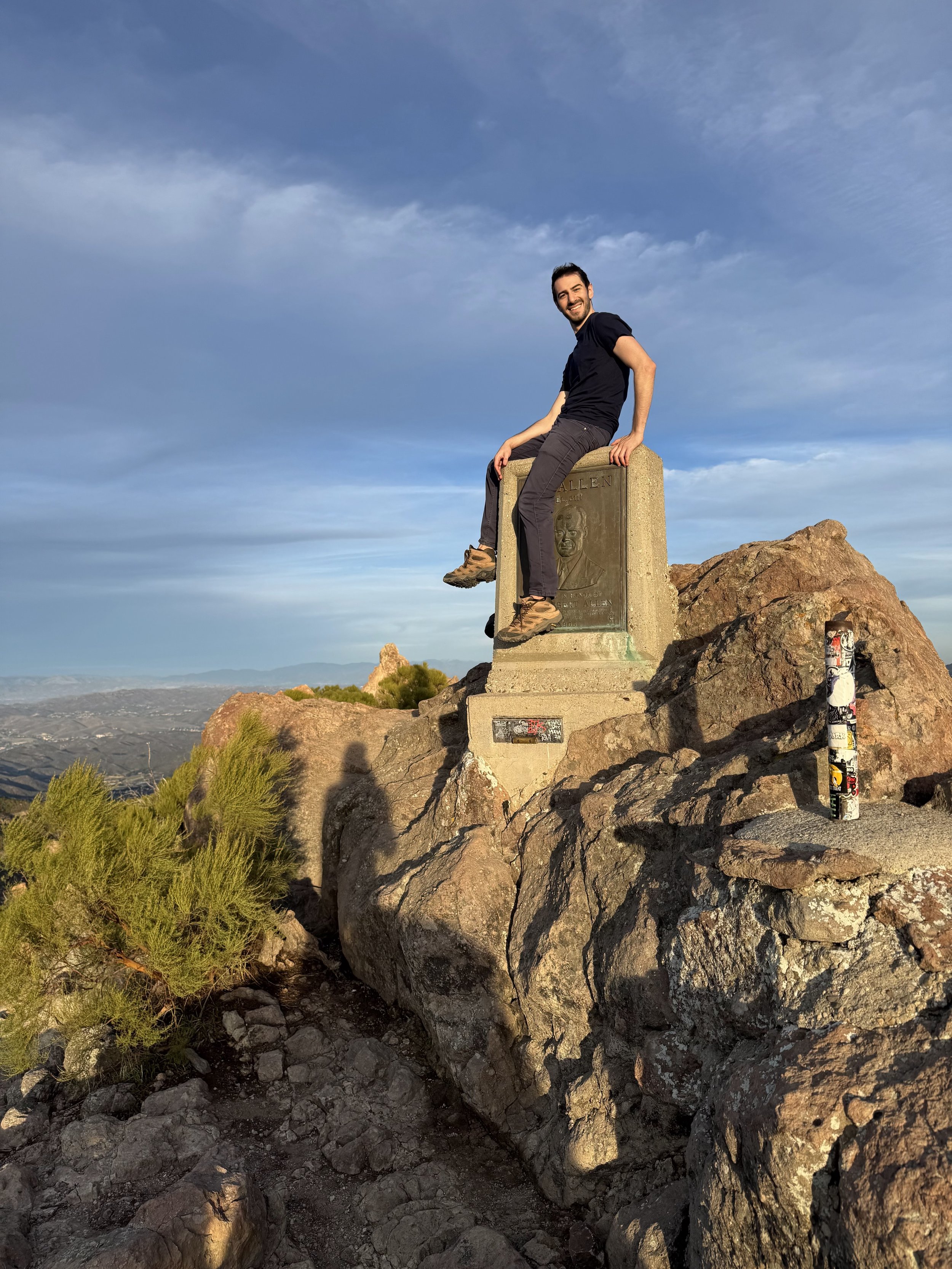 A man sitting on a memorial plaque atop a rocky hill, with an expansive view of the landscape and sky in the background.
