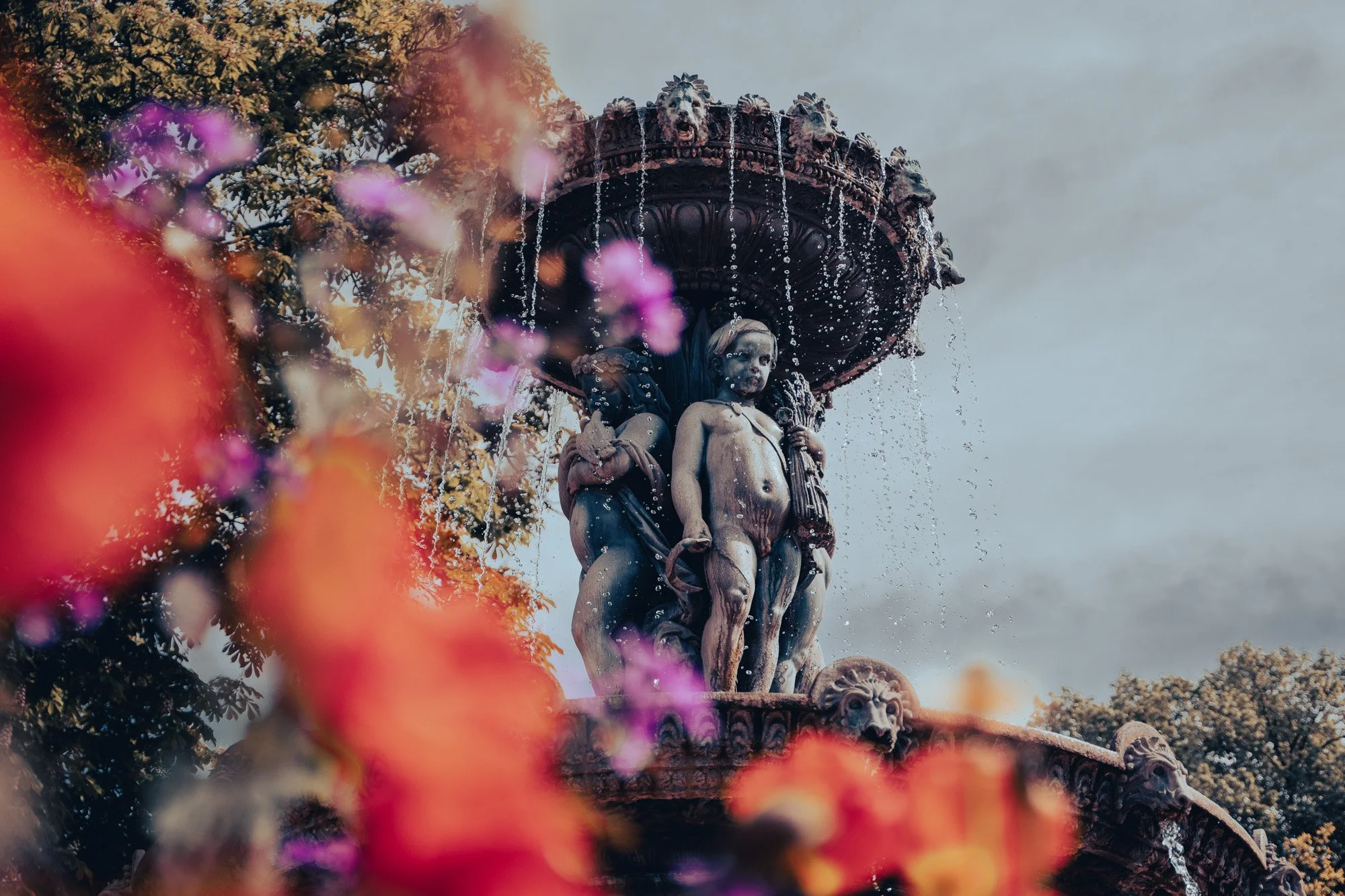 A detailed ornamental fountain with sculpted figures and water cascading from the top, partially obscured by blooming flowers in the foreground.