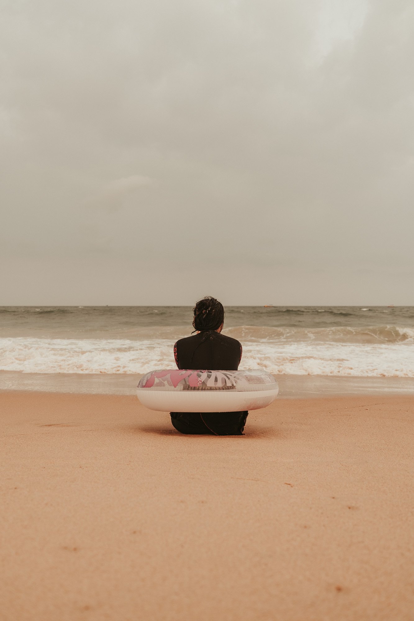 A person sitting on the beach facing the ocean with a float ring around their waist.