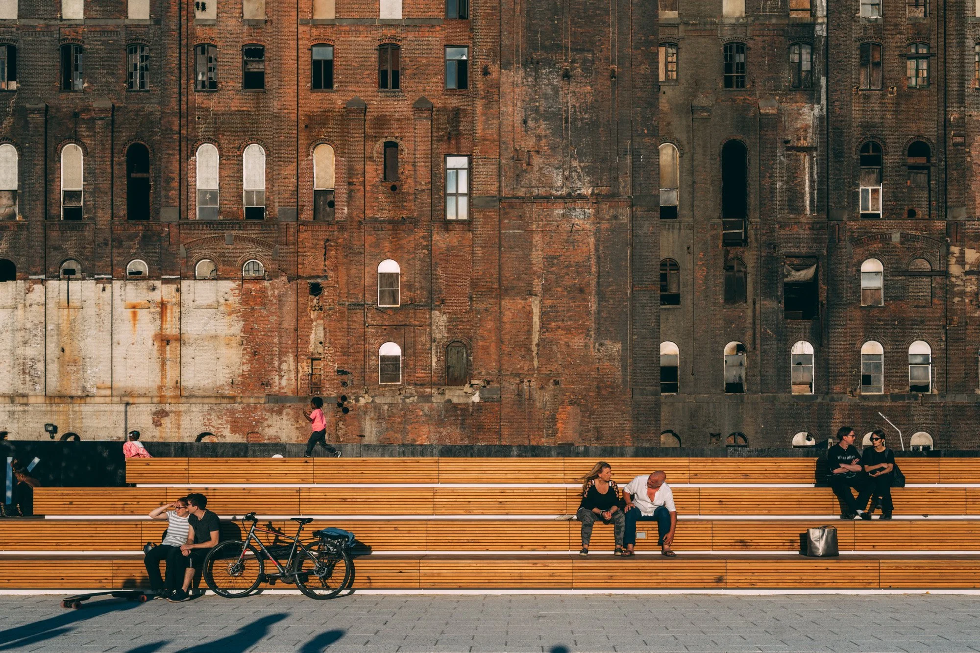 People sitting on wooden benches and walking in front of a large old brick building with many windows.
