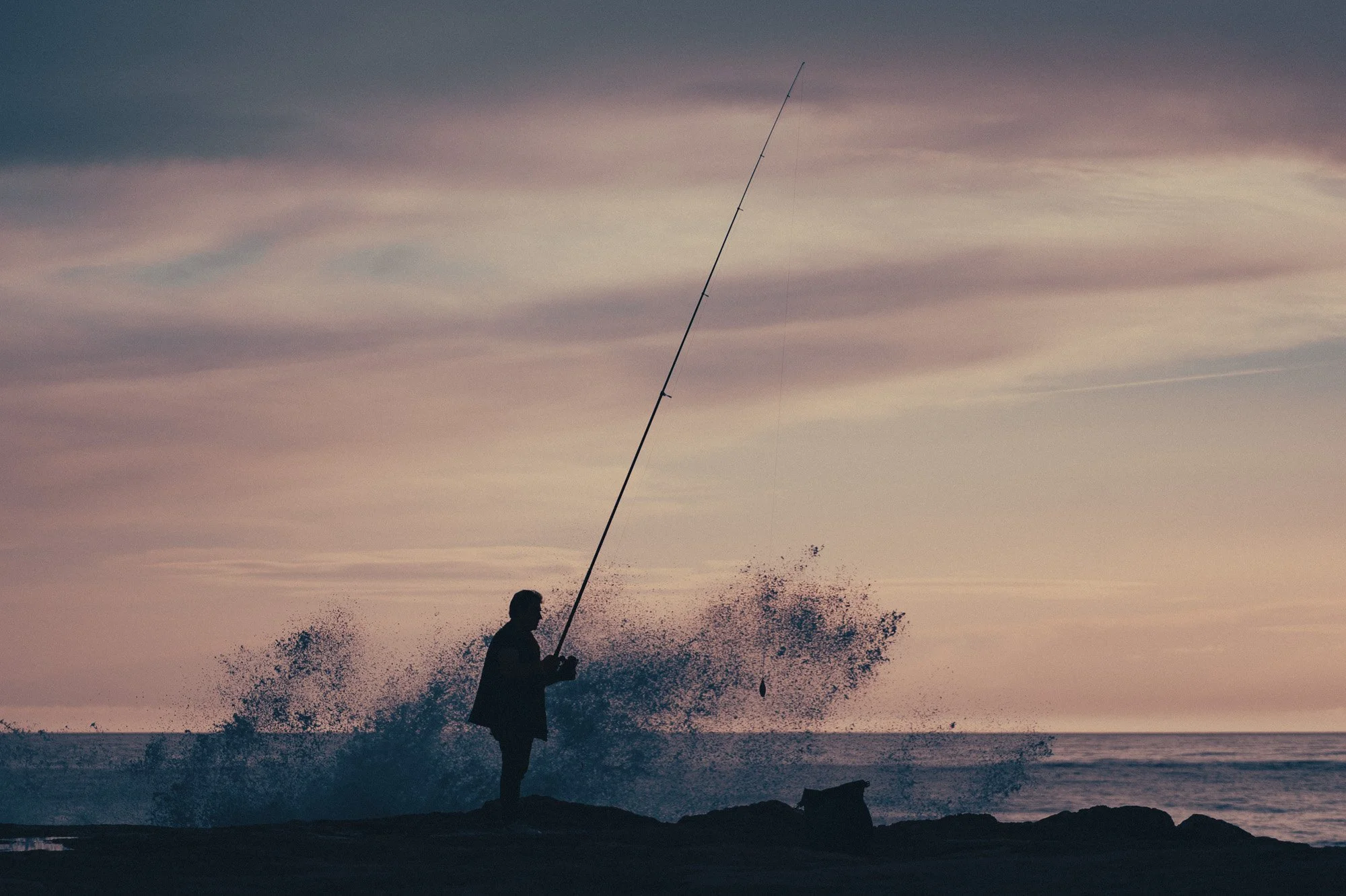 Person fishing by the ocean during sunset, with waves crashing and a cloudy sky.