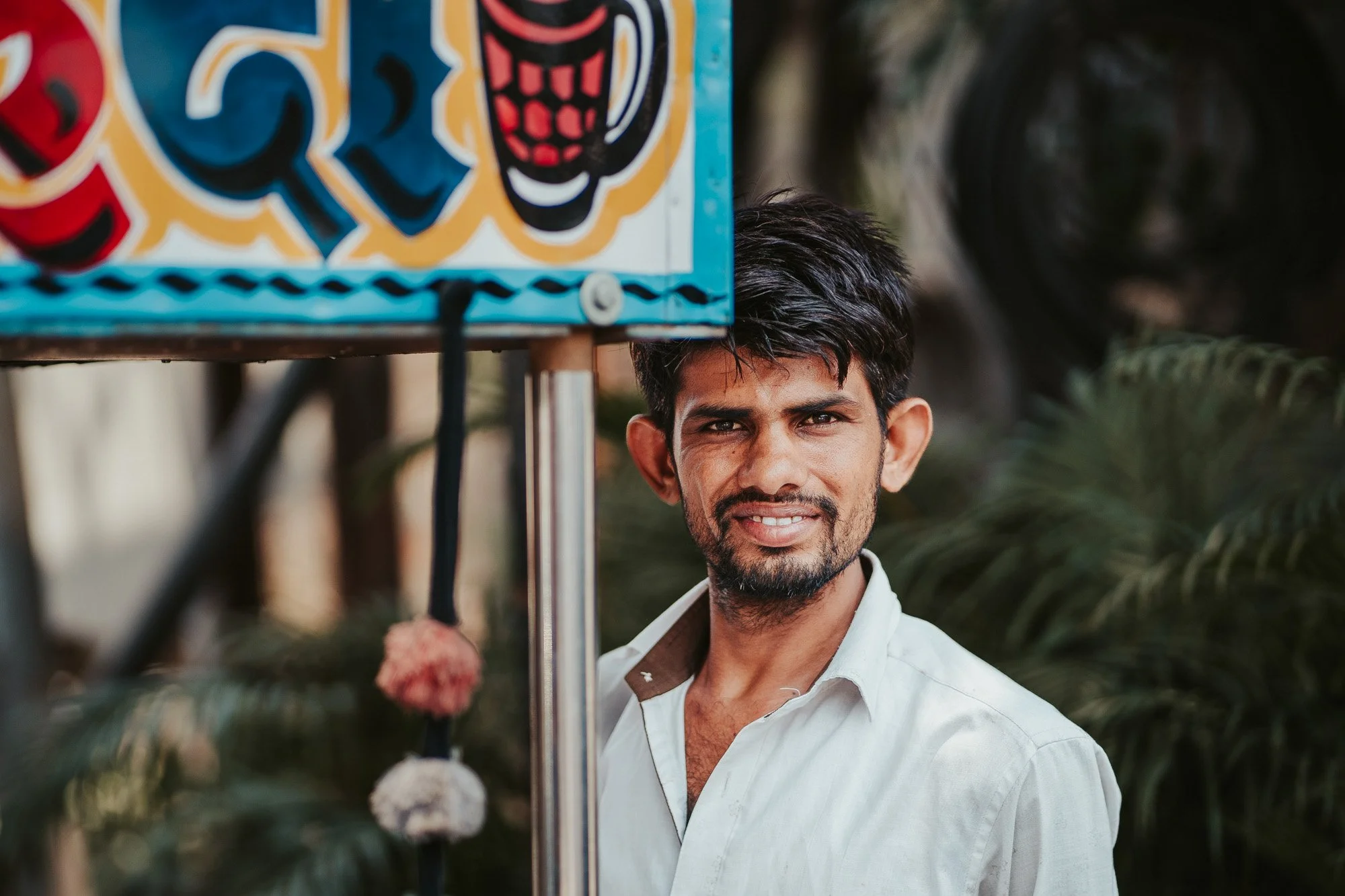 A man with dark hair and beard smiling in front of a colorful sign, with greenery in the background.