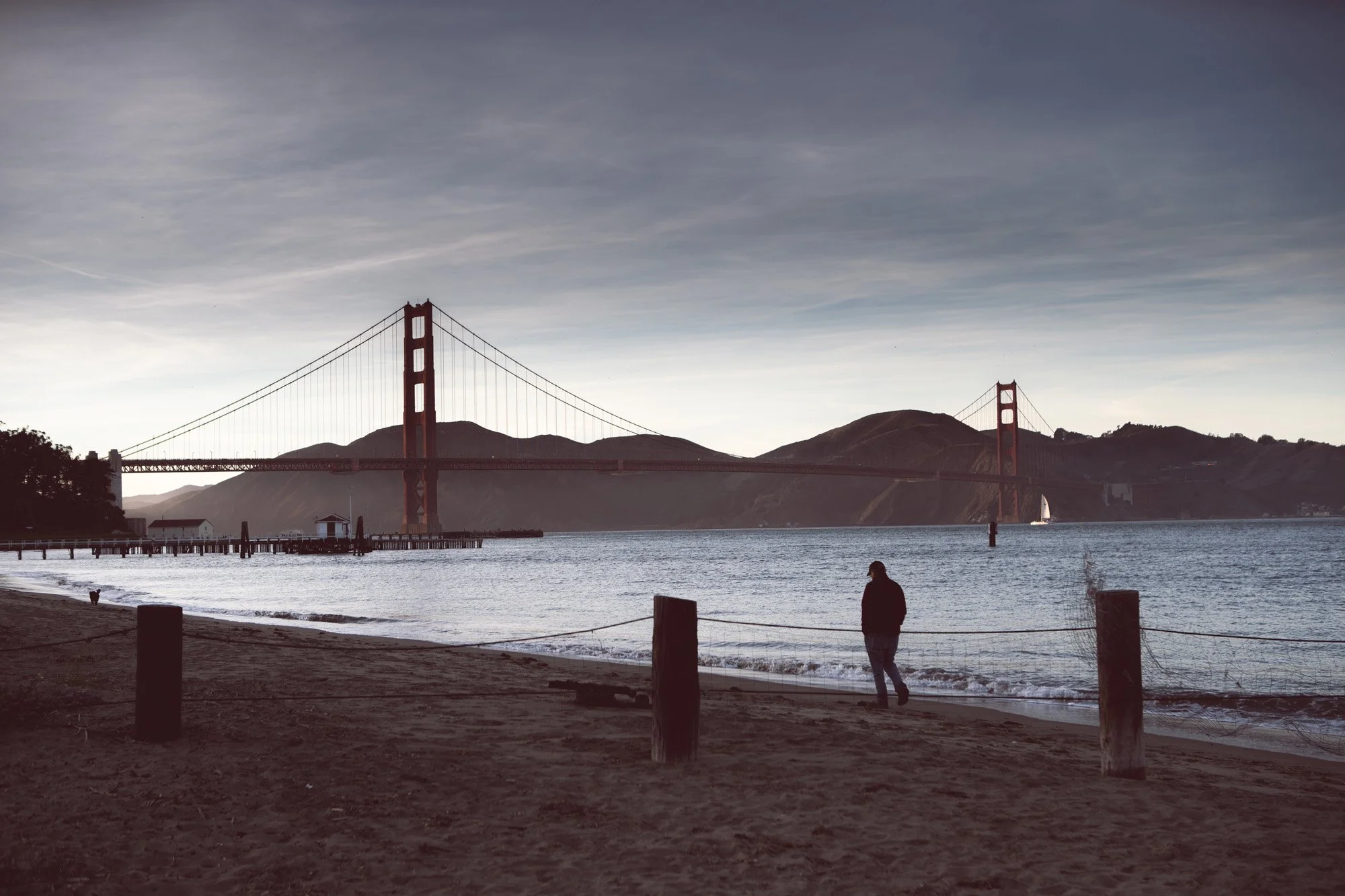 A person walking along a beach with the Golden Gate Bridge in the background during sunset or sunrise.