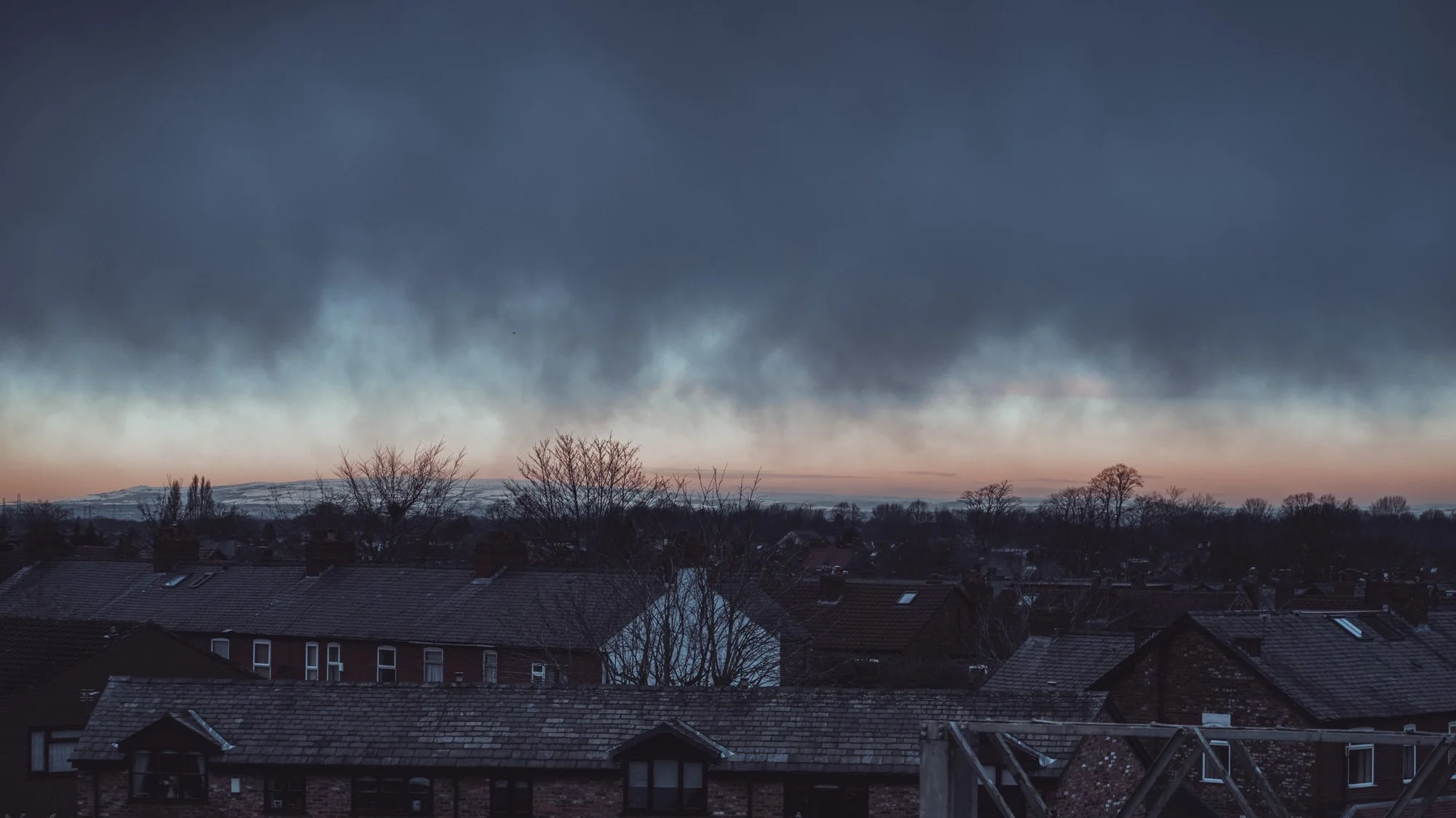 The image shows a dark, cloudy sky over a neighborhood with rooftops and leafless trees at dusk, with hills in the background.