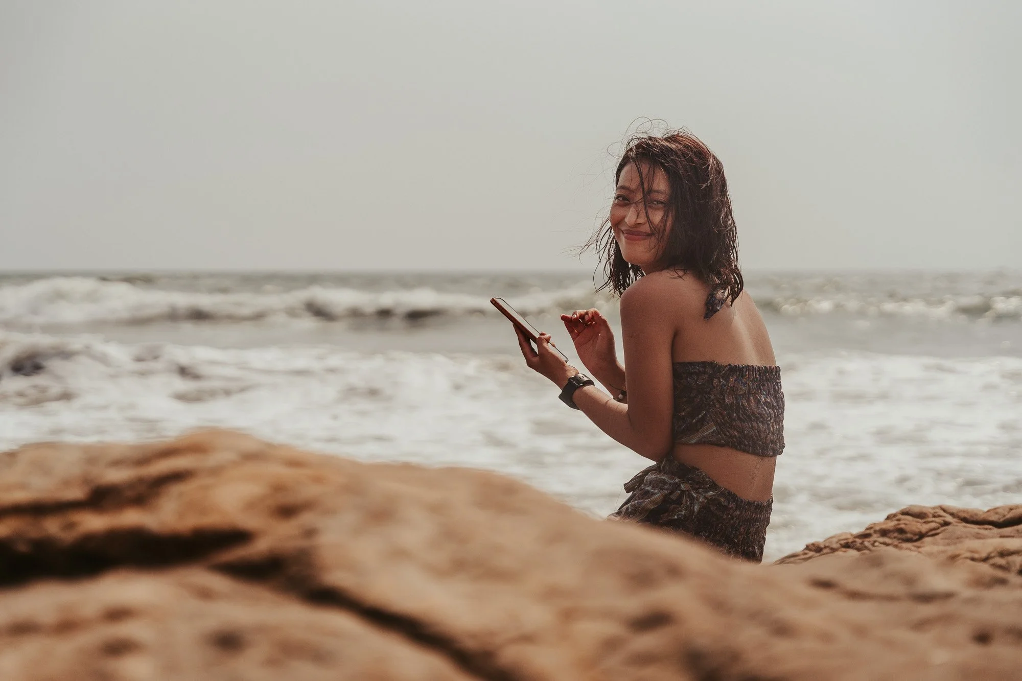 Young woman sitting on rocks at the beach, smiling while looking at her phone.