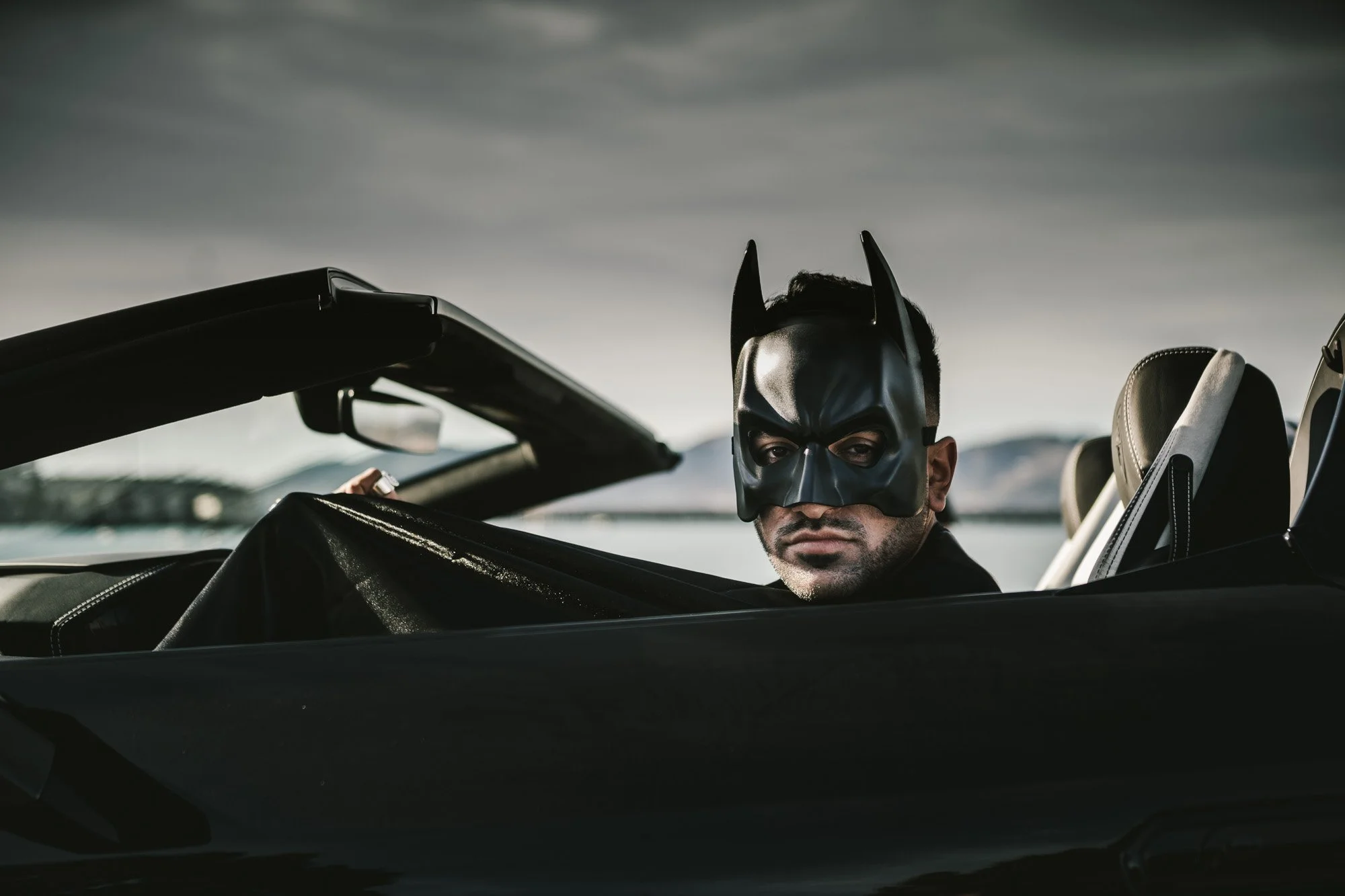 Man wearing a Batman mask sitting in a black convertible car, looking at the camera with a serious expression, against a cloudy sky and a distant landscape.
