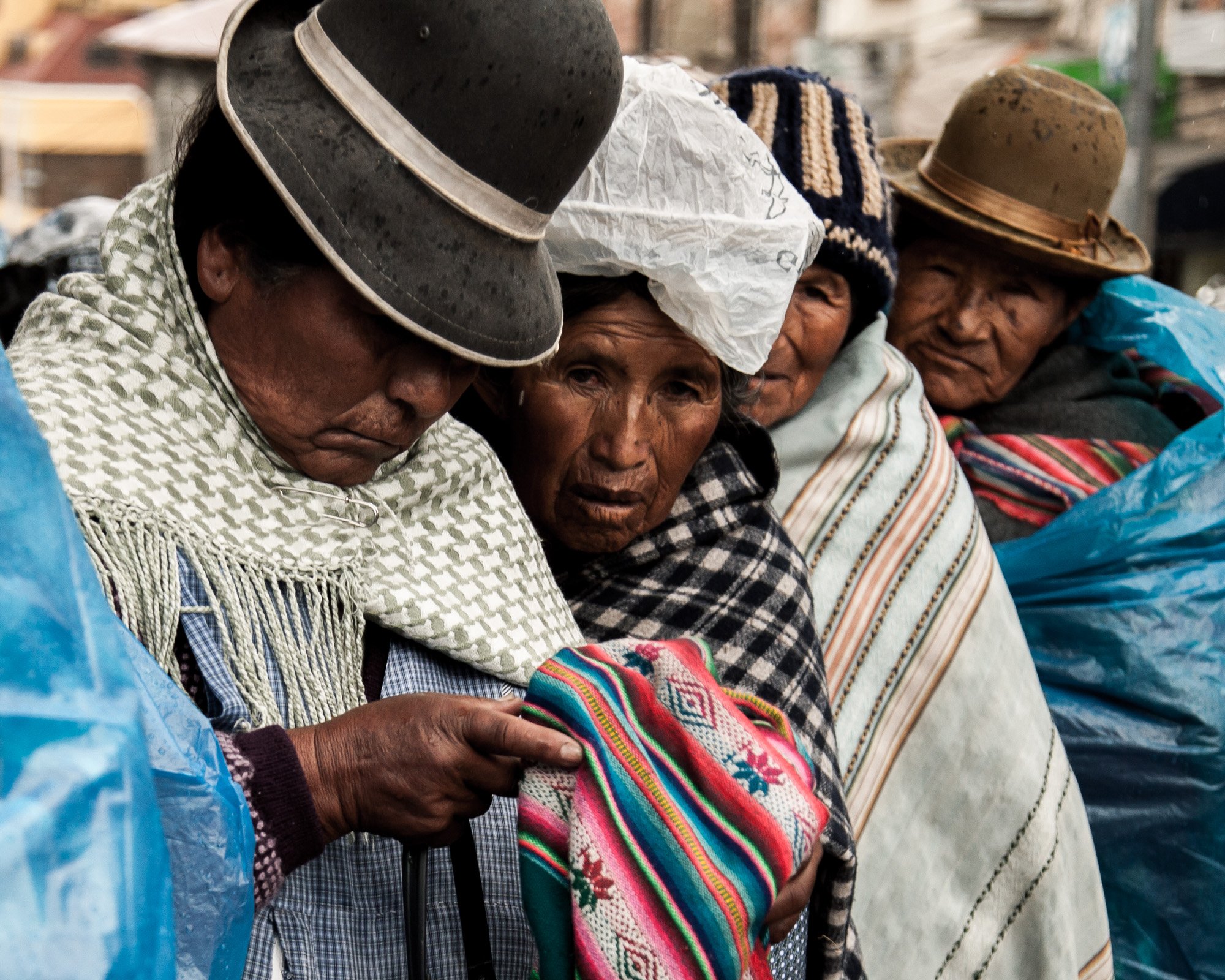 Group of indigenous women wearing traditional clothes and hats, holding colorful textiles in a market setting.