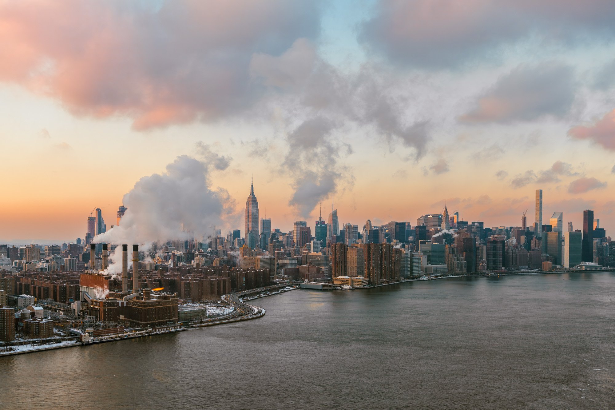 Skyline of New York City with boats on the river, Empire State Building in the center, and smoke rising from factory chimneys at sunset.