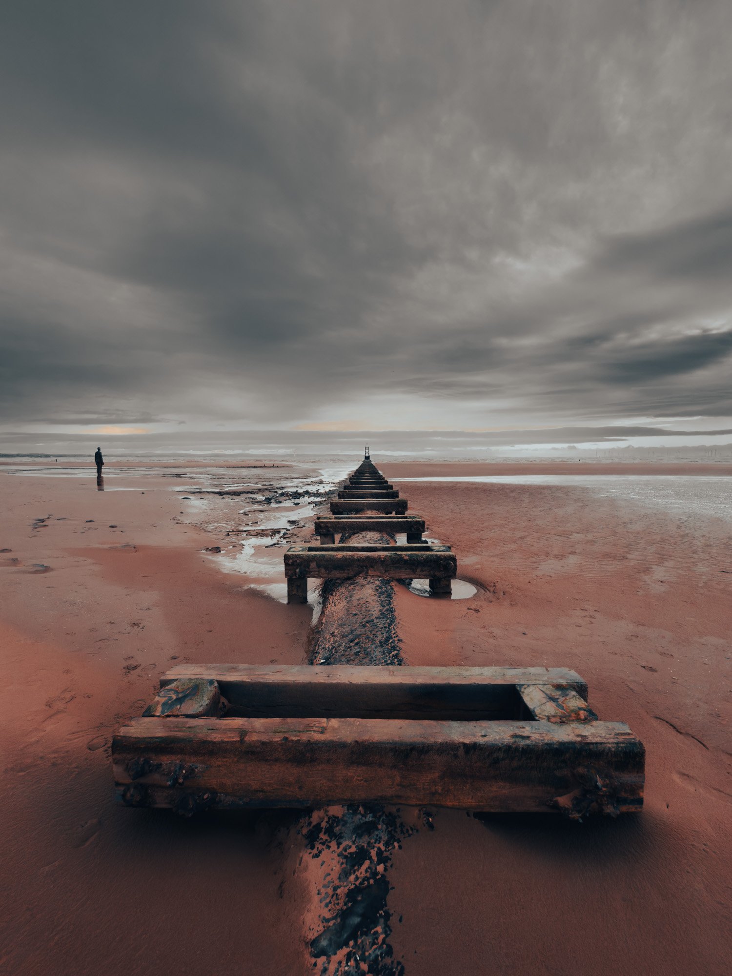 A rusty pipe extending from the sandy beach out to the horizon under a cloudy sky, with a lone person standing on the beach.