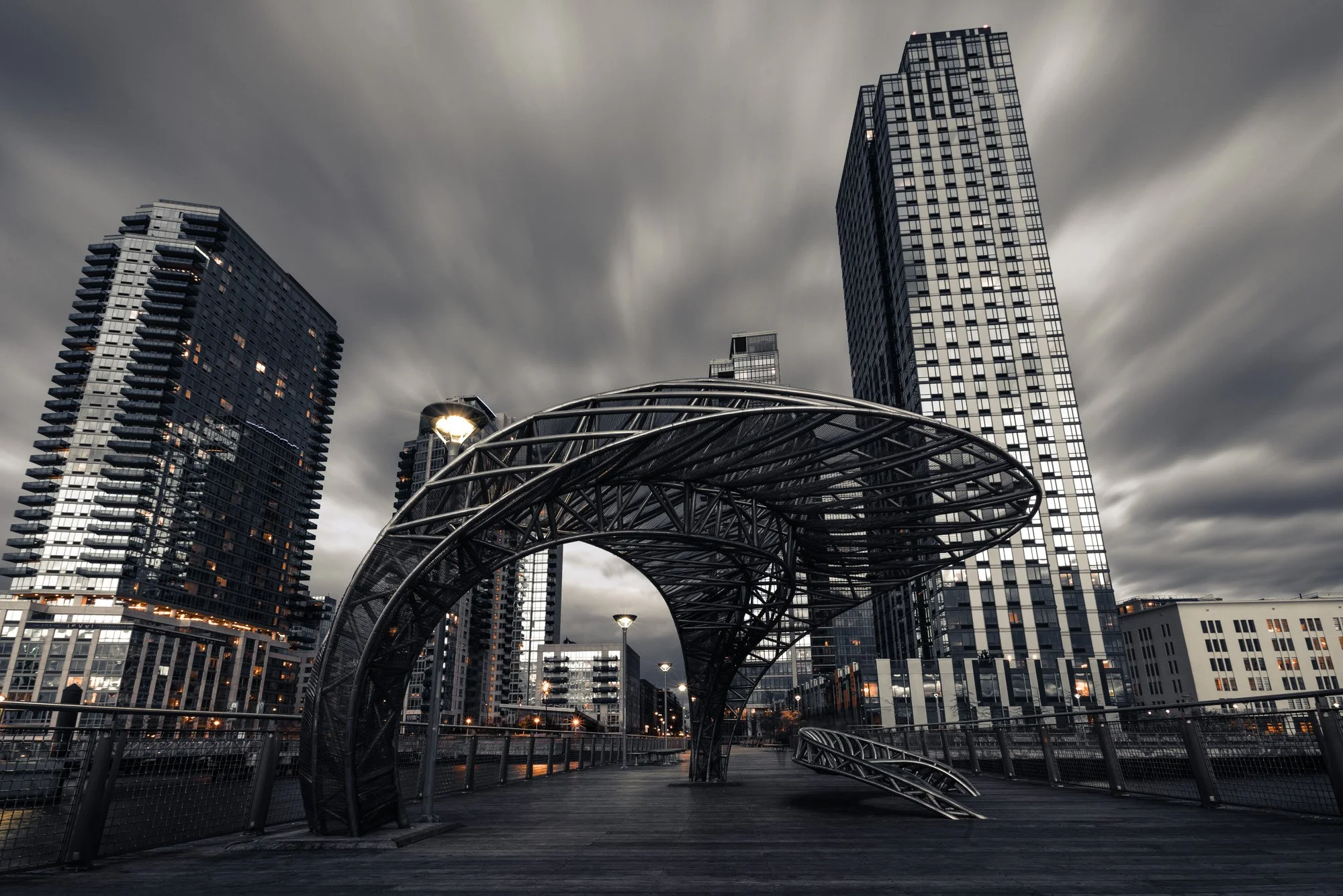 A modern cityscape featuring tall skyscrapers with reflective glass windows against a cloudy, overcast sky. In the foreground, a curved metal architectural structure is present on a wooden boardwalk with streetlights along the path.