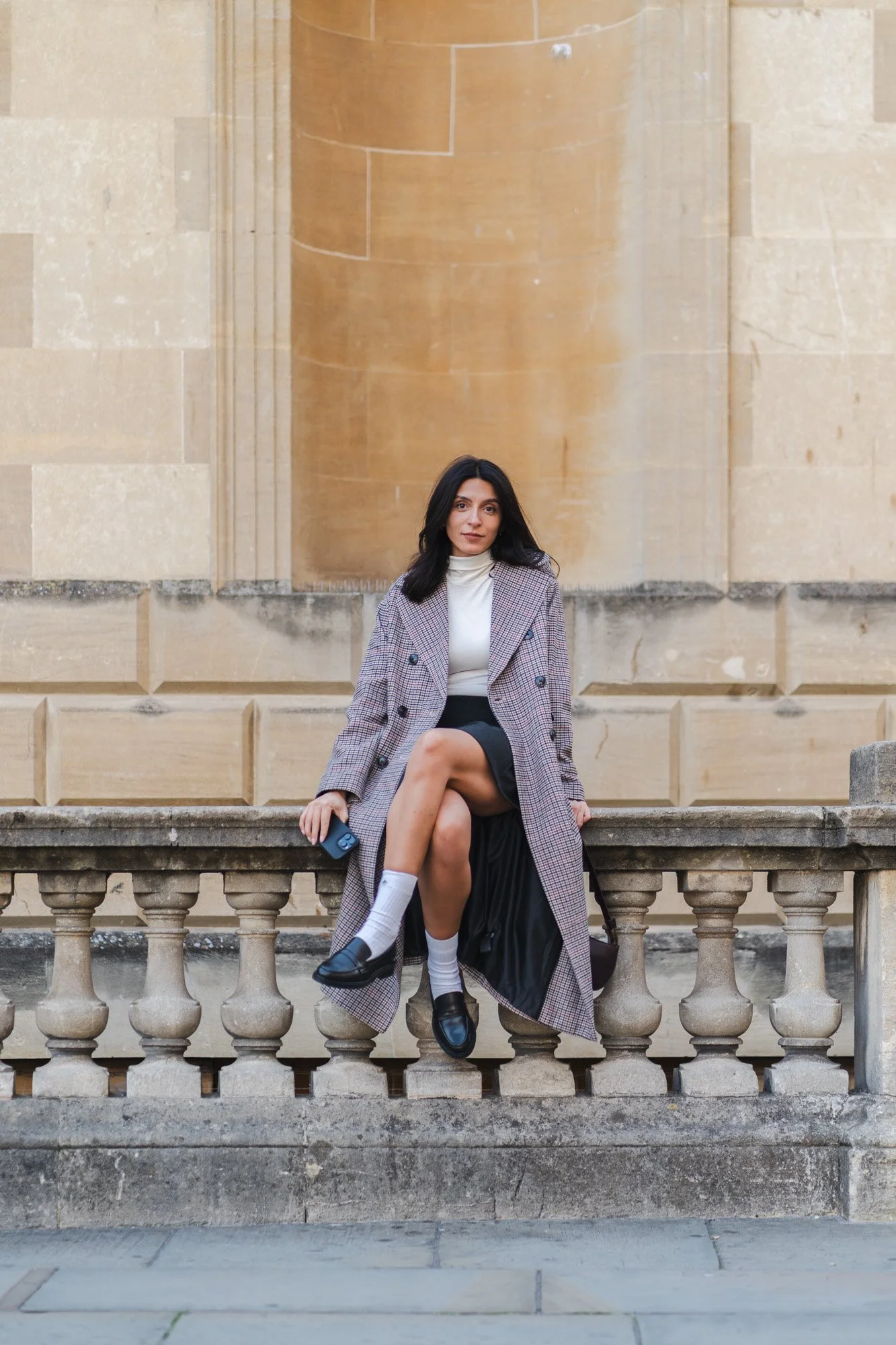 A woman sitting on a stone railing in front of a beige building, wearing a gray checkered coat, white turtleneck, black skirt, white socks, and black loafers, holding a phone.