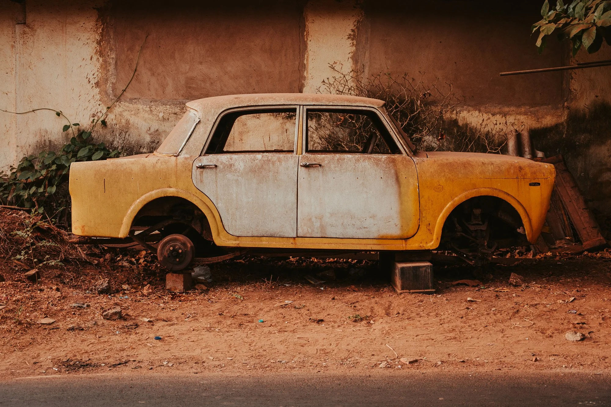 An abandoned, weathered yellow and white car missing its wheels, propped up on blocks, and sitting on dirt ground against a worn wall.