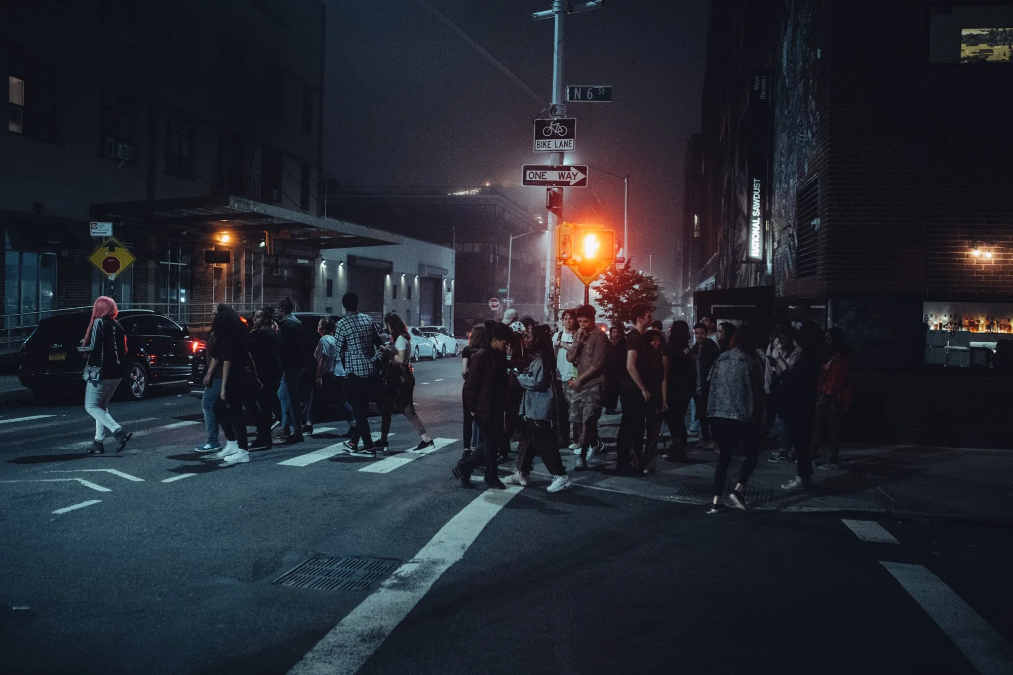 A group of people crossing a city street at night, illuminated by street lights and a traffic signal, with cars parked along the curb and a building with signage in the background.