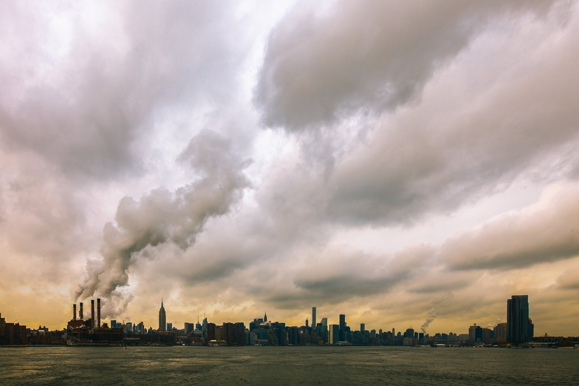 City skyline with tall buildings under a cloudy sky and smoke or steam rising from factories.