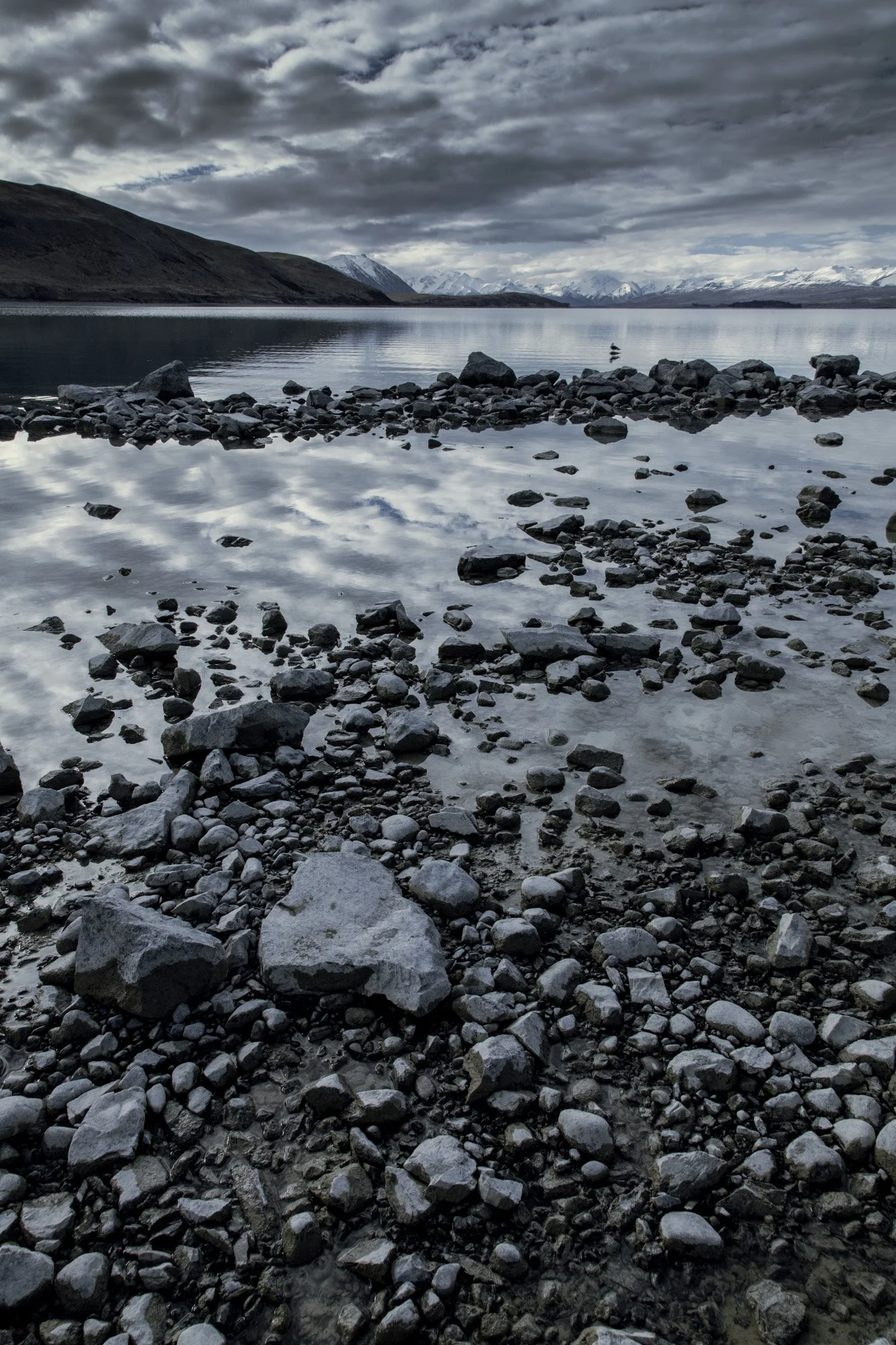 A rocky lakeshore under a cloudy sky, with mountains in the distance.