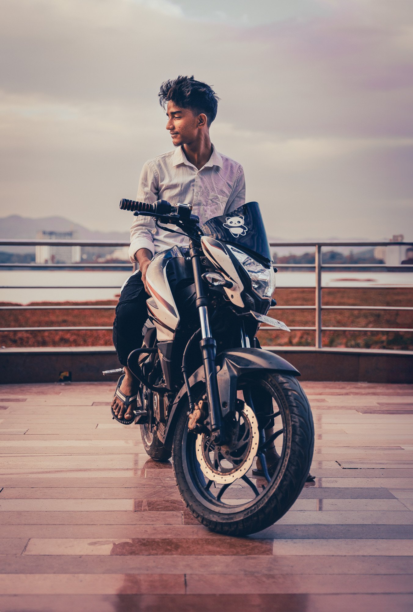 Young man in a white shirt and dark pants sitting on a black and white motorcycle on a rooftop with a city view and cloudy sky in the background.