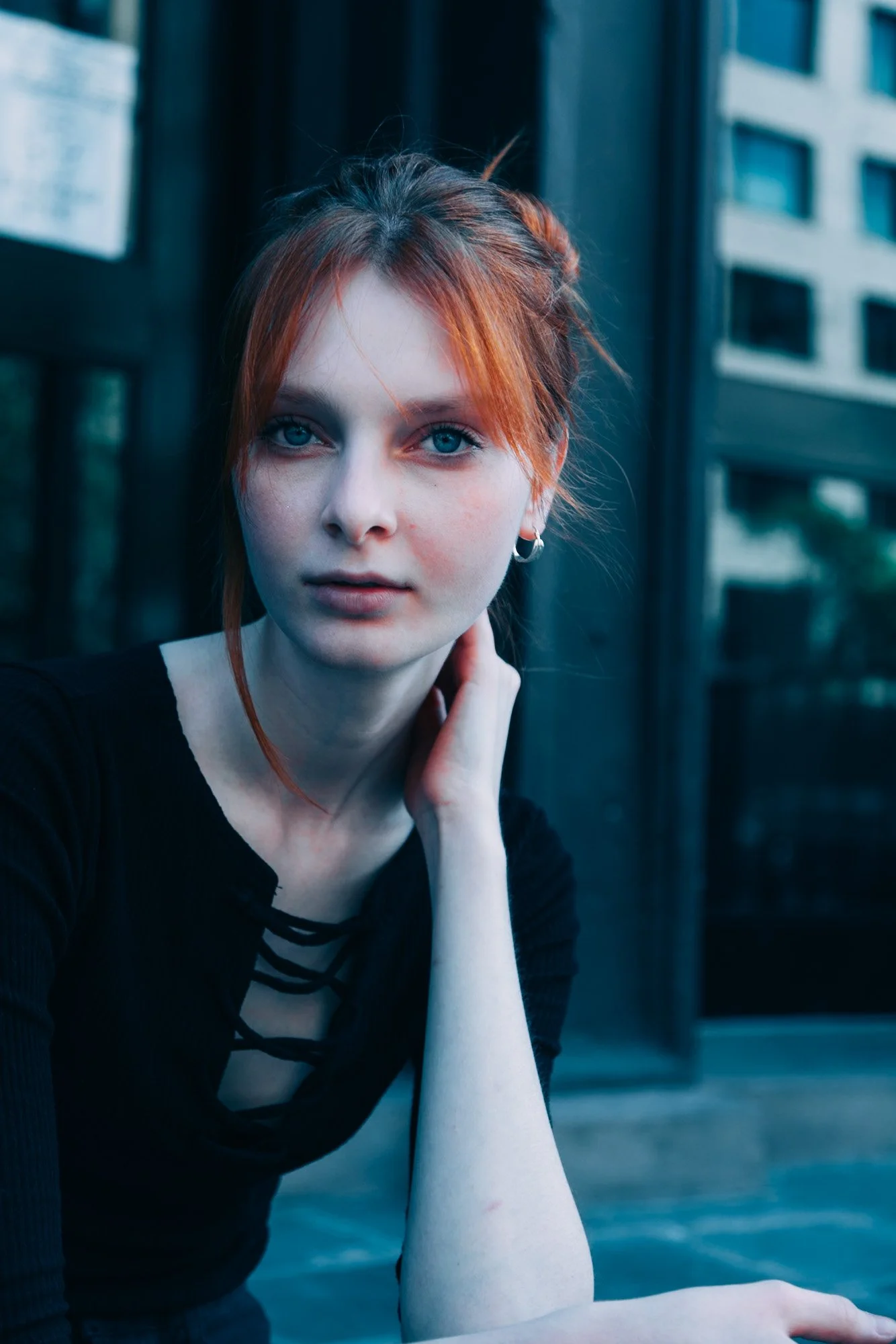 Close-up of a young woman with red hair and blue eyes, wearing a black top with laced-up front, sitting outdoors near a glass building.