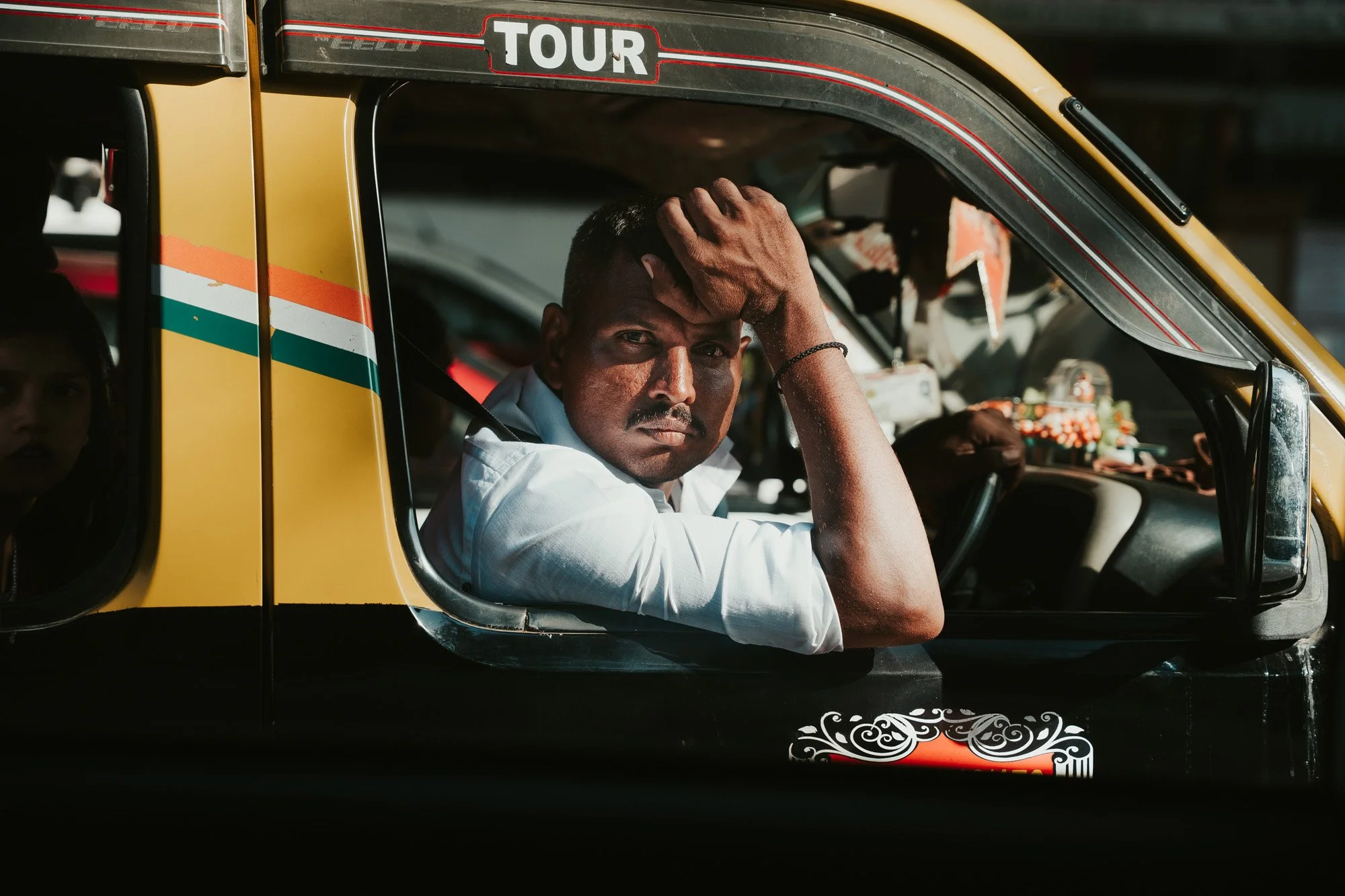A man sitting in the driver's seat of a taxi with his arm resting on the window frame, looking directly at the camera with a serious expression.