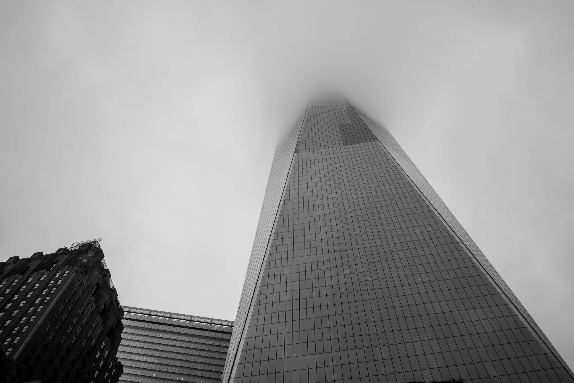 A tall skyscraper with glass panels reaching into cloudy, foggy sky, viewed from below.
