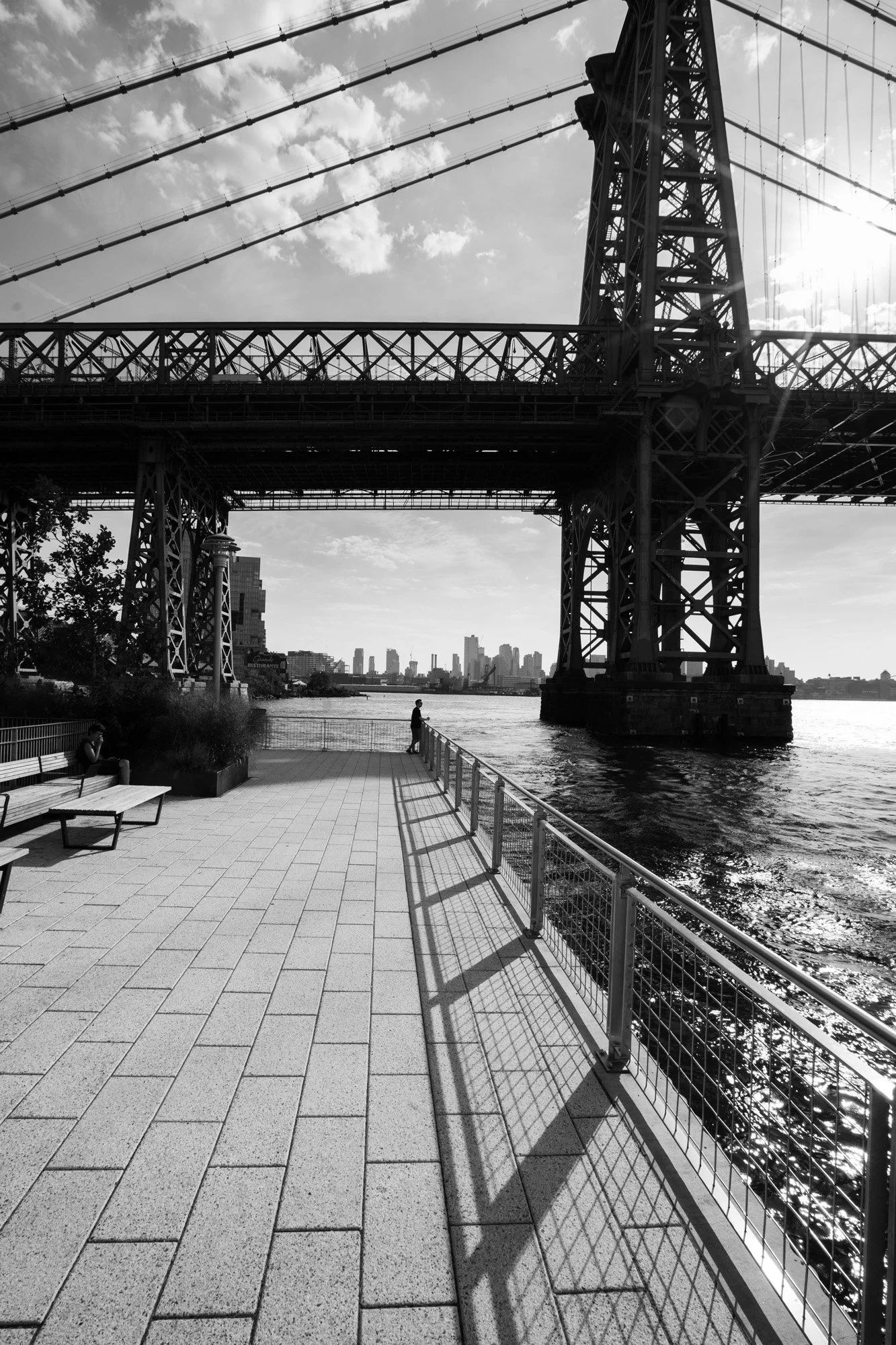 A black and white photo of a city waterfront with a large bridge overhead, a person standing on the pier near the water, and a city skyline in the background.