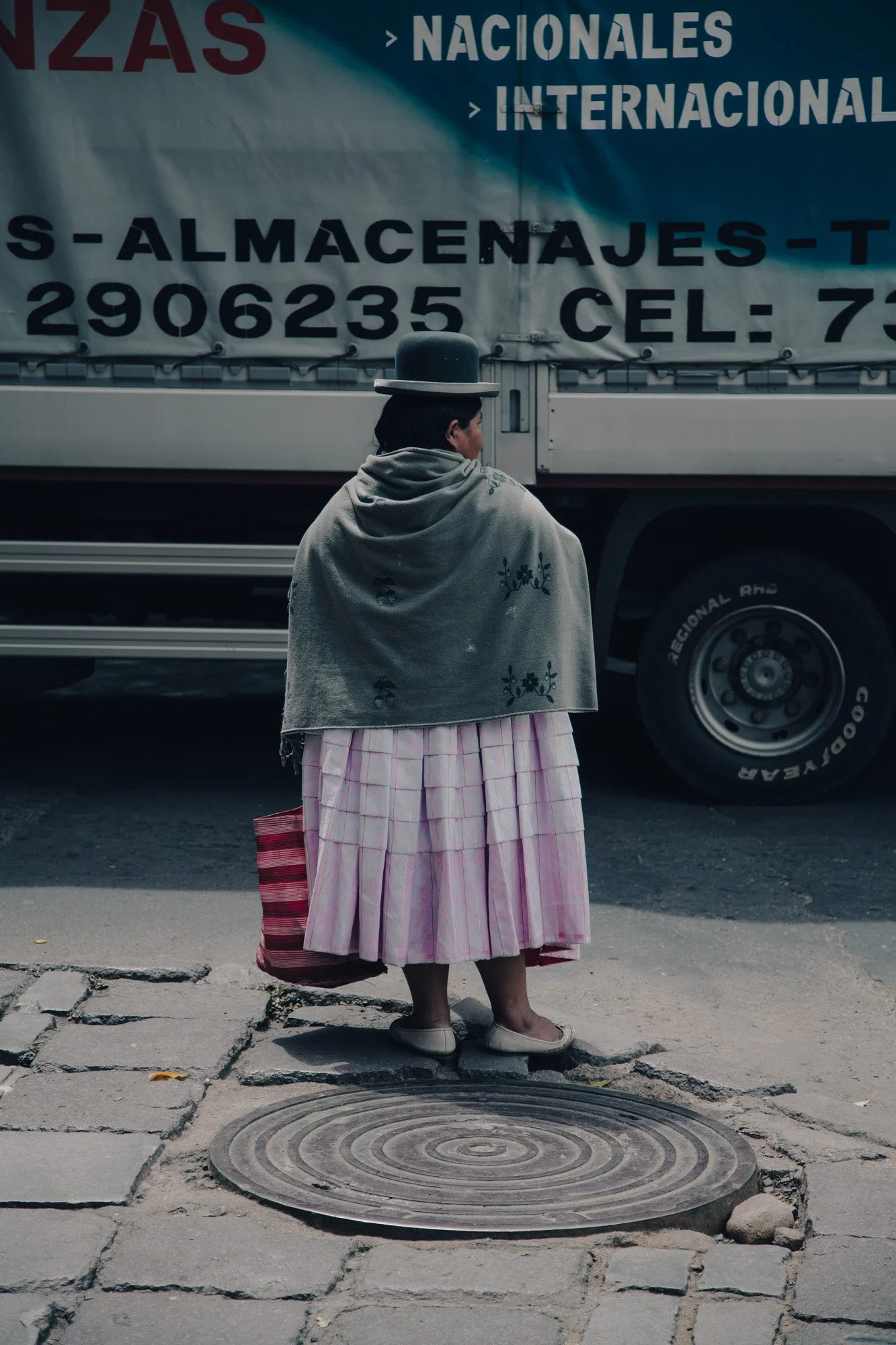 A woman dressed in traditional clothing, including a gray shawl, pink skirt, and a hat, holding a red and pink striped bag, standing on a city sidewalk near a parked truck.