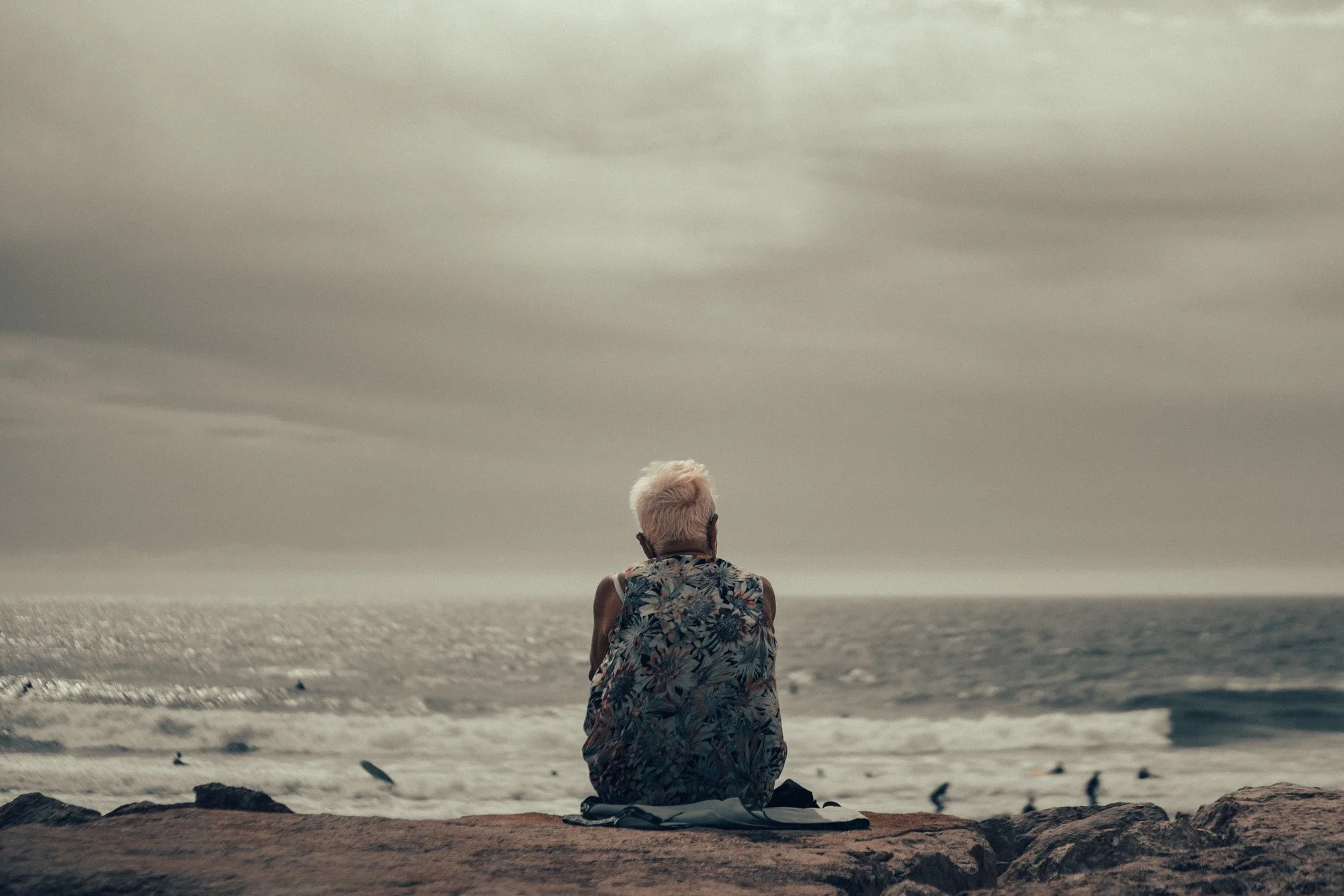 A person with light-colored hair sitting on a rock by the ocean, facing the water and cloudy sky.