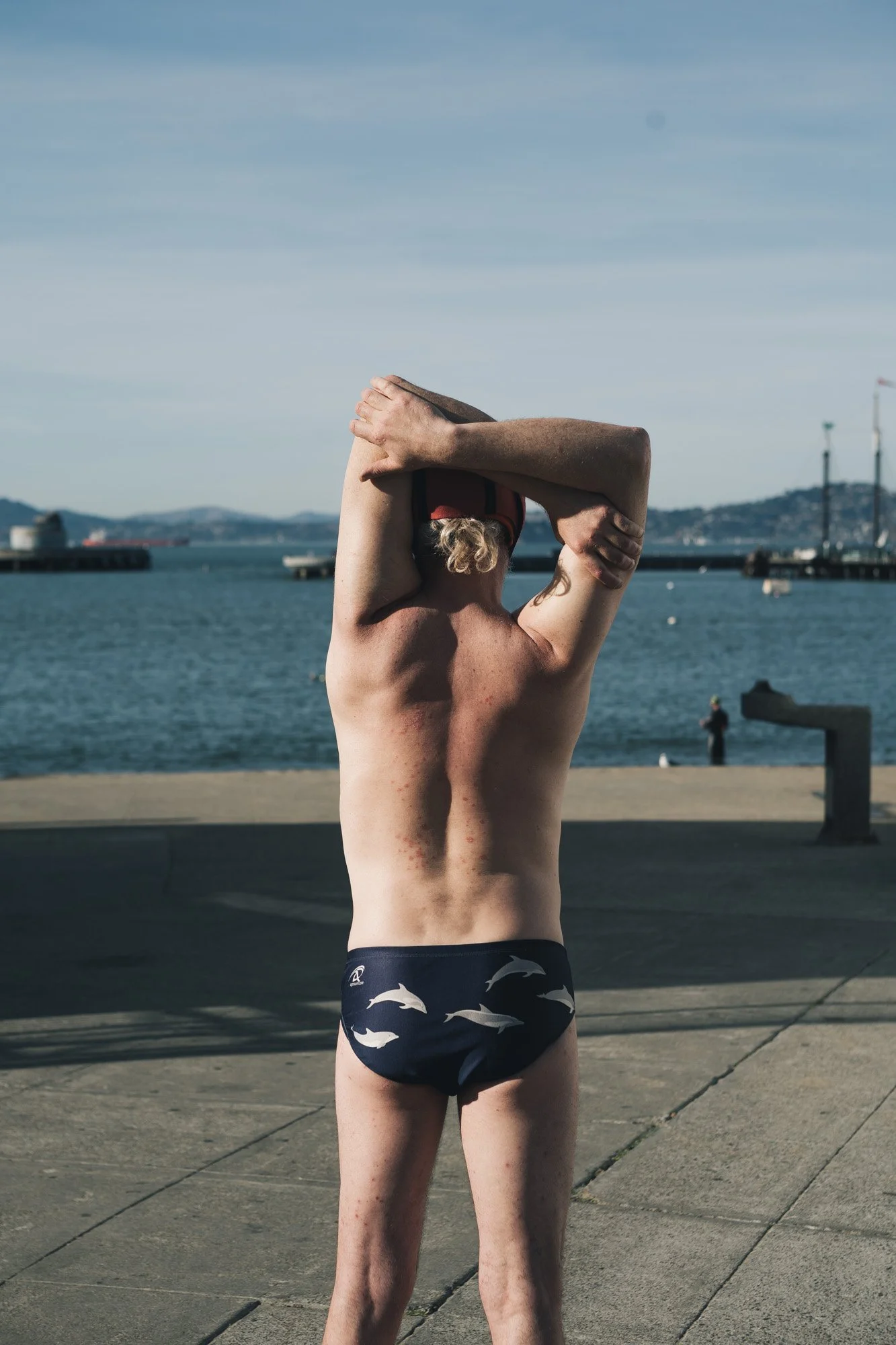 A young man standing on a waterfront promenade with his back to the camera, wearing swimming trunks with dolphin prints, stretching with arms behind his head, near a body of water with islands and cranes in the distance.