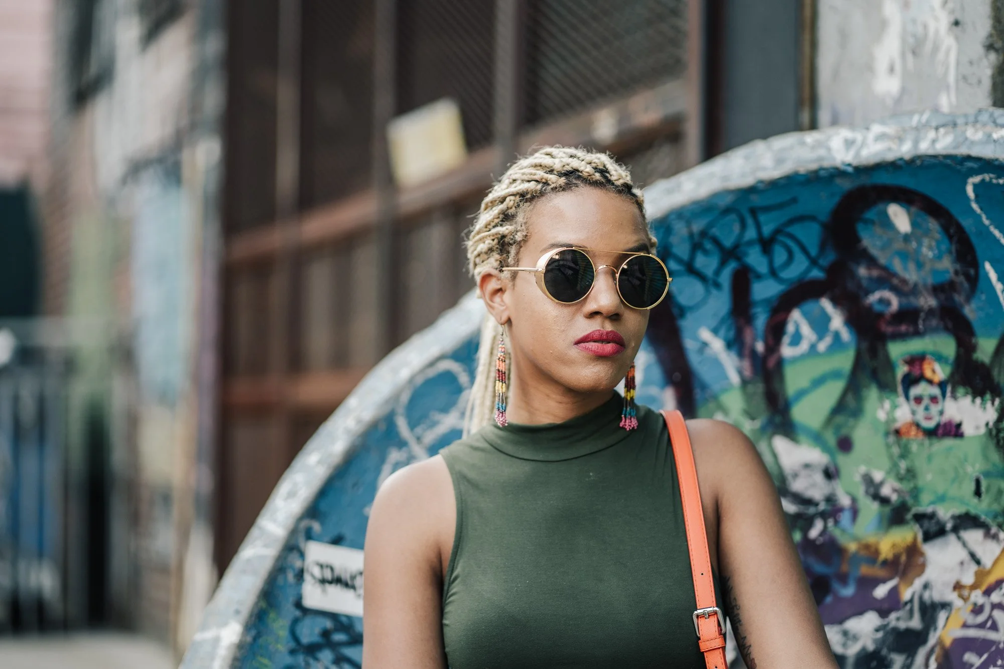 A stylish woman with blonde twisted hair, wearing round sunglasses and colorful earrings, stands in front of a graffitied skate ramp. She is dressed in a sleeveless green top and carrying an orange strap purse.