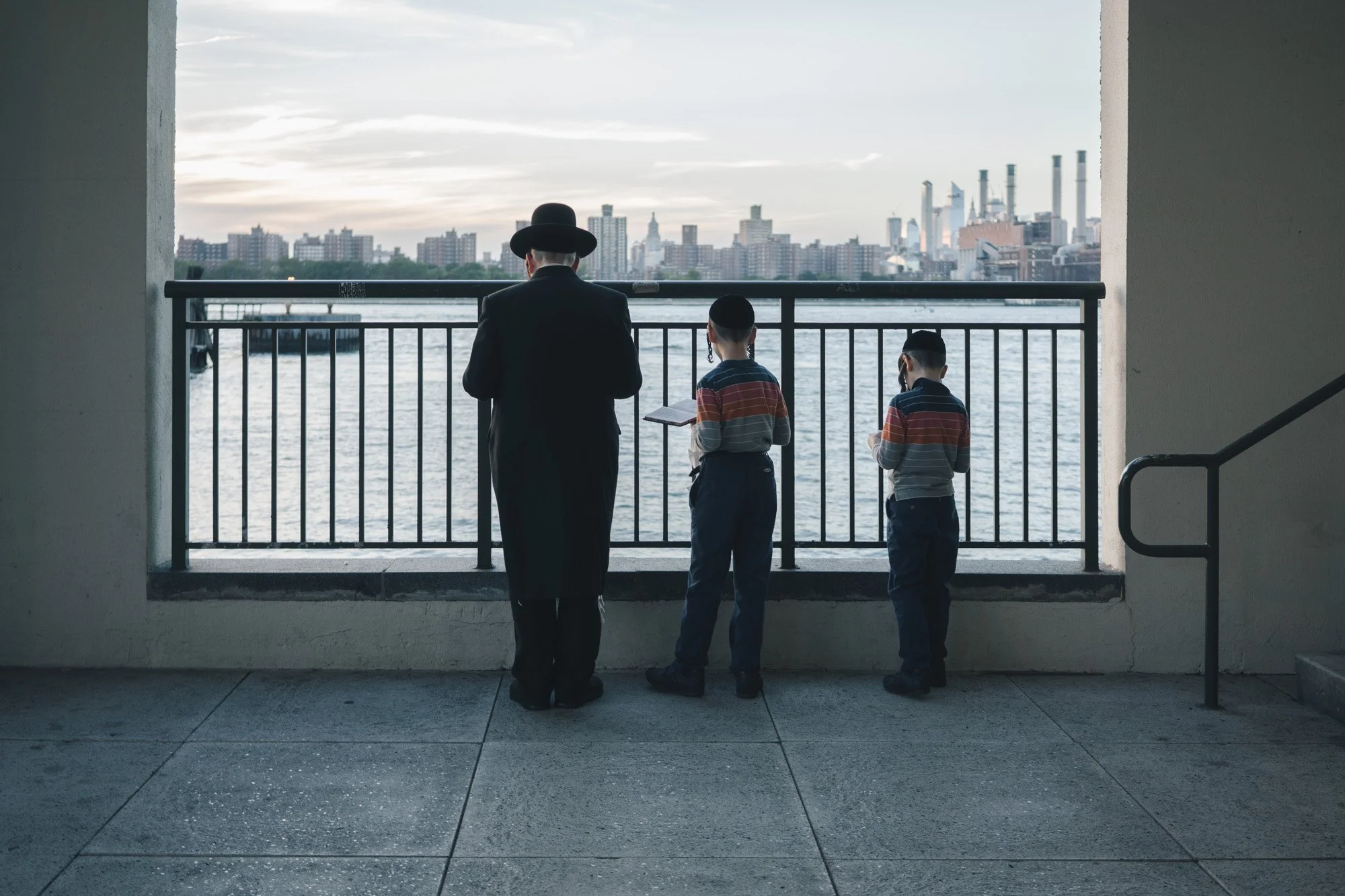 An adult male and two boys standing on a balcony overlooking a city skyline with water in the foreground, during dusk.