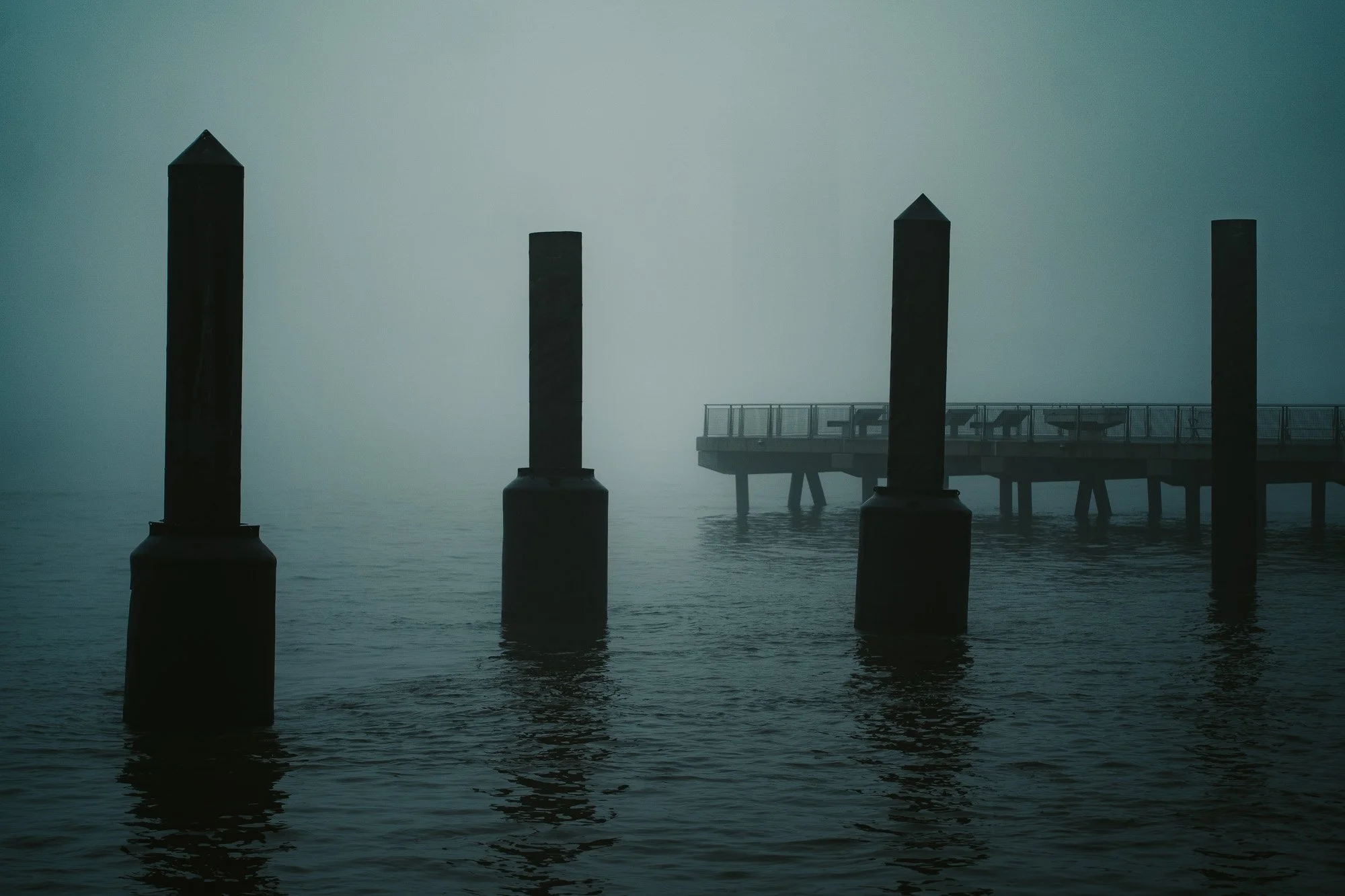 A foggy scene over water with four dark pilings and a pier in the background.