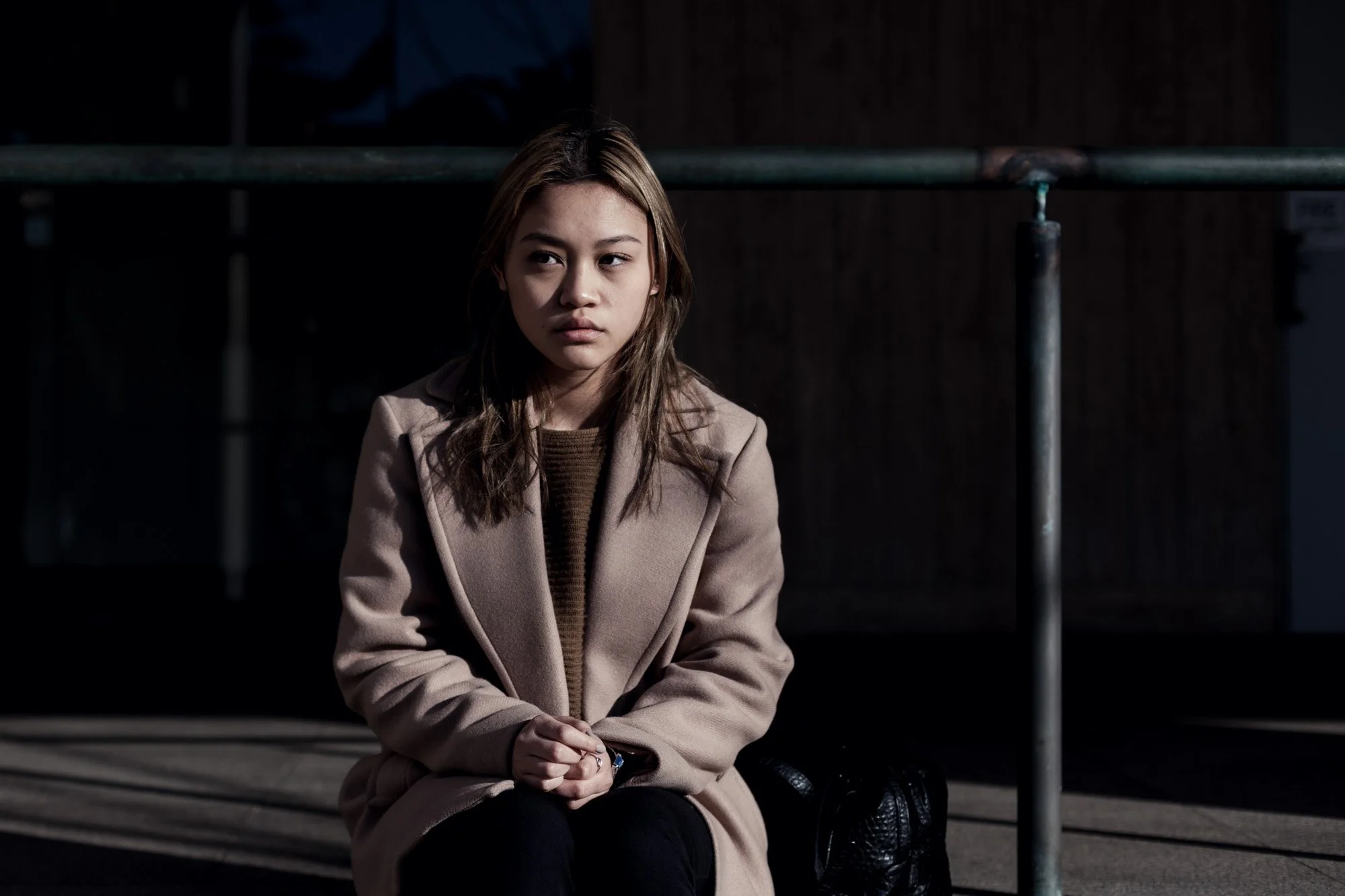 A young woman with light brown hair, wearing a beige coat, sits on a bench in a dimly lit environment, looking to her left with a thoughtful expression.