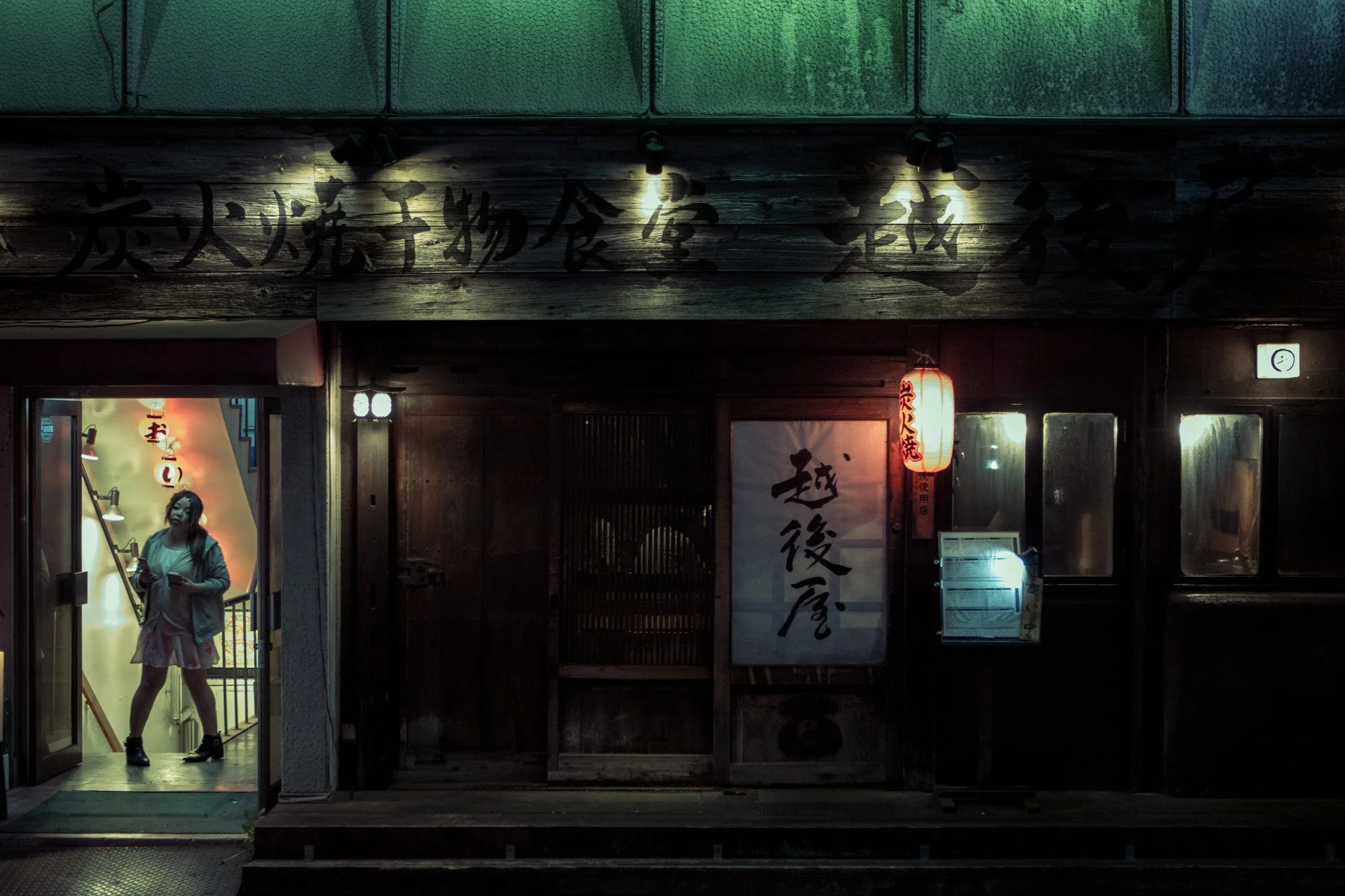A woman standing at the entrance of a Japanese restaurant with traditional wooden signage and red paper lanterns outside, evening setting.